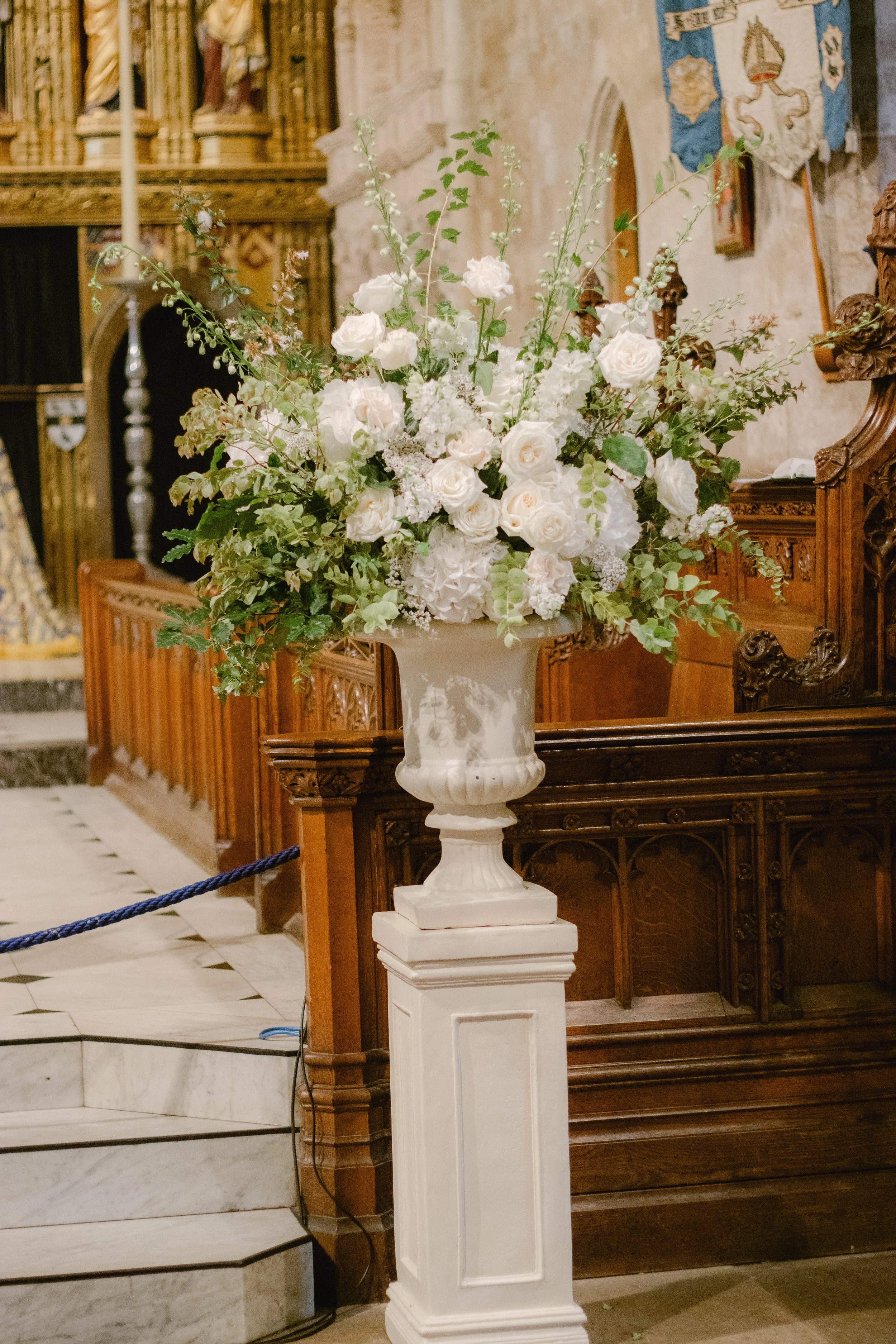 A large white floral arrangement with roses and greenery in a white pedestal urn inside a church.
