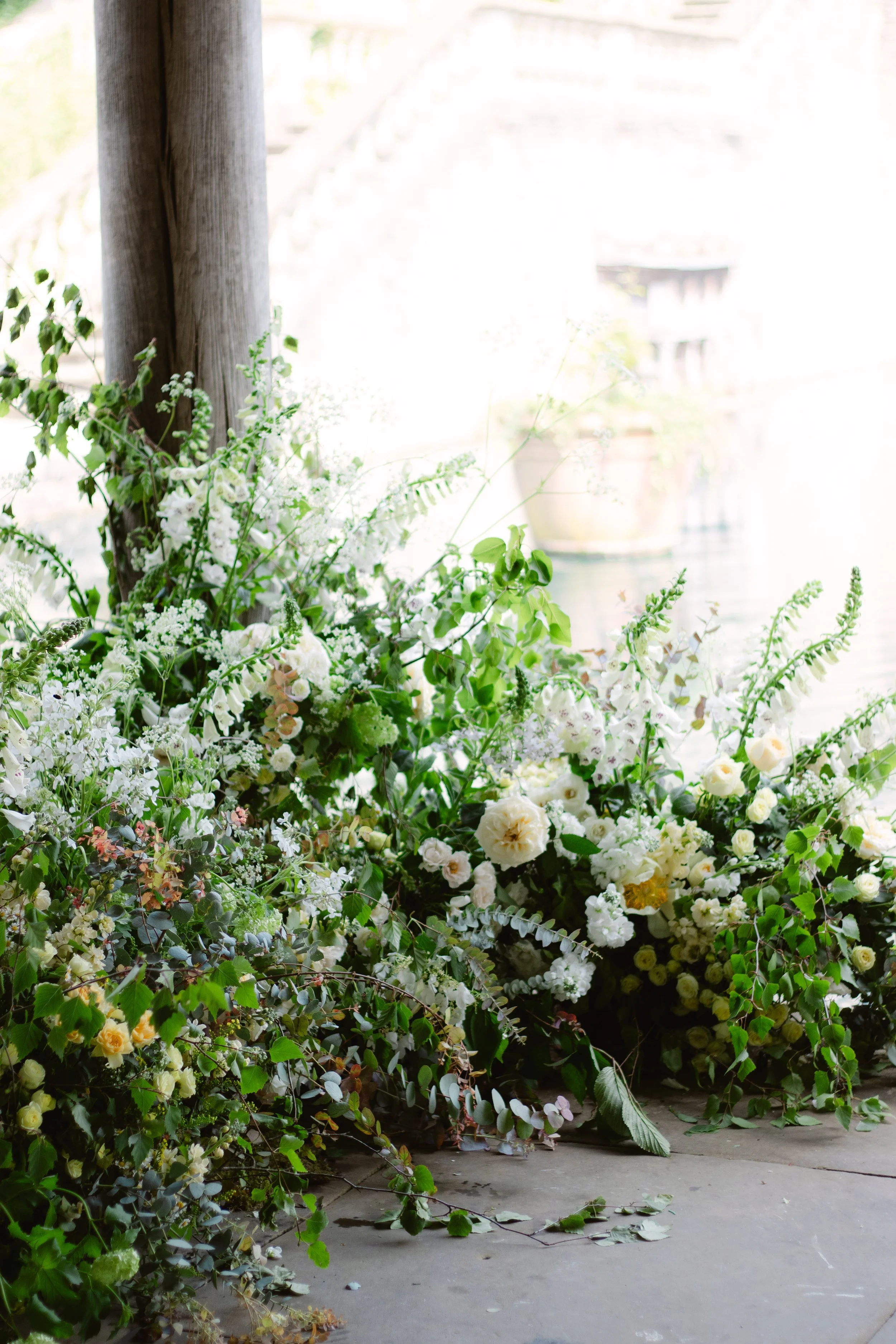 Arranged white and green floral display with roses, ferns, and other greenery near a wooden post, against a bright background.