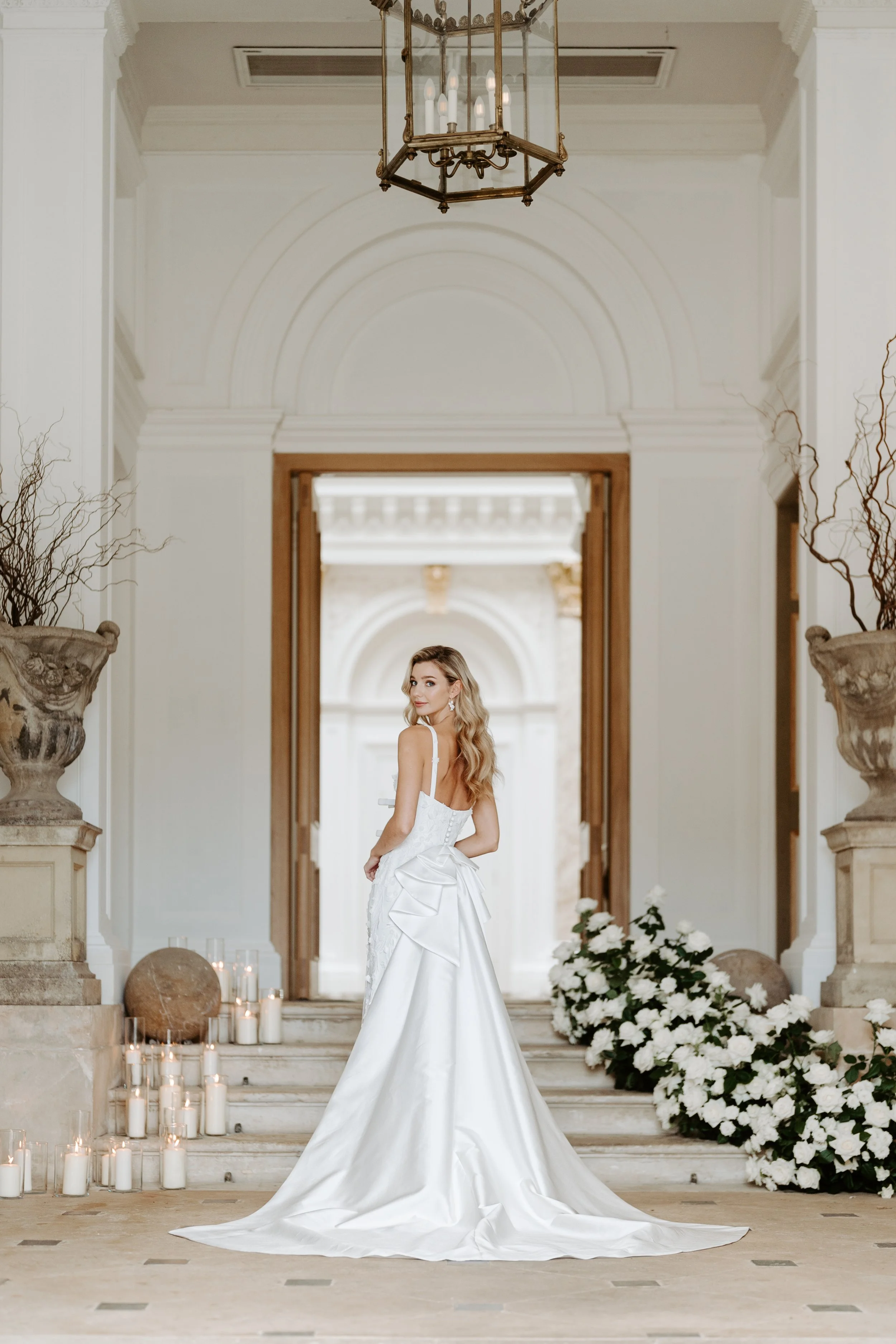 A woman in a white wedding gown stands on a staircase in an elegant, classical-style interior with tall white walls, decorative columns, candles, and floral arrangements.