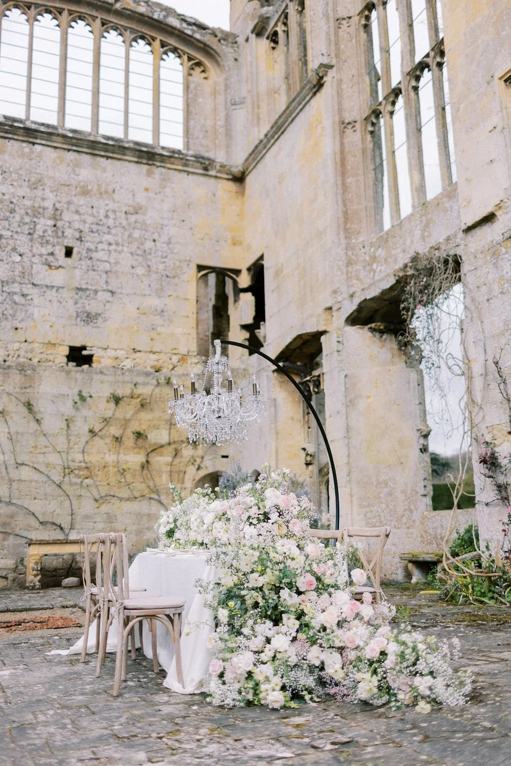 An elegant wedding or event setup with a round table covered with a white tablecloth, adorned with a large floral arrangement of white and light pink flowers, and decorated with a crystal chandelier hanging above. The setting is in front of an old, p
