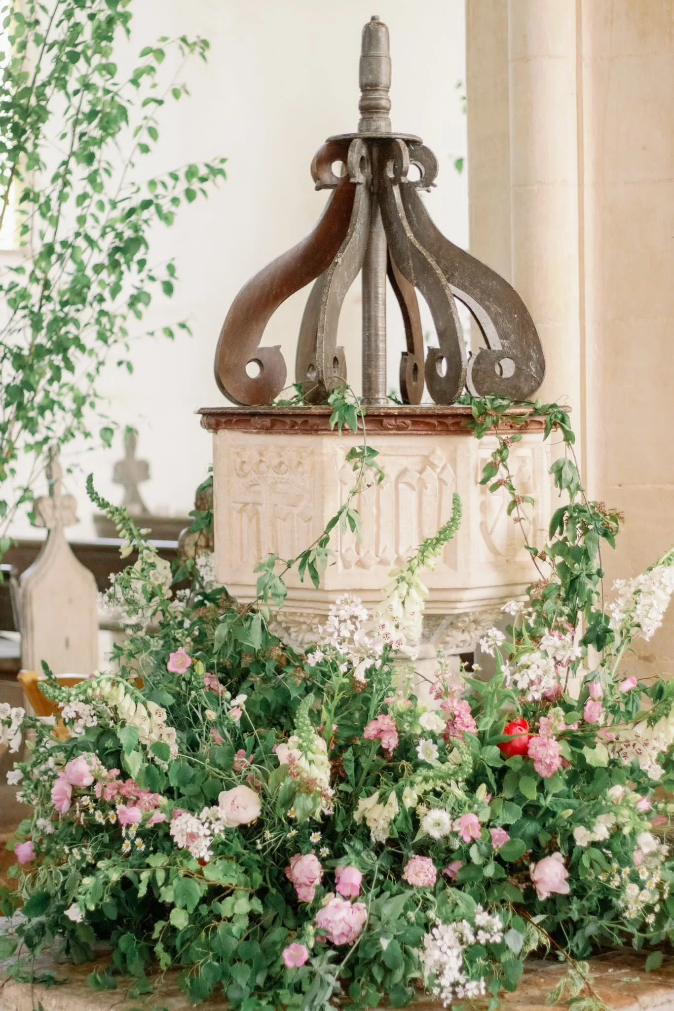 A vintage well or fountain with a shephard's hook structure on top, surrounding a carved stone or plaster base, adorned with lush pink and white flowers and greenery at the base.