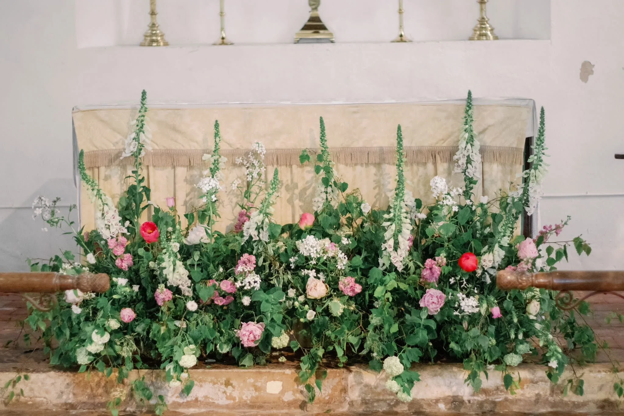 Arrangement of pink and white flowers with green foliage on a stone ledge in front of a decorated altar with candlesticks.