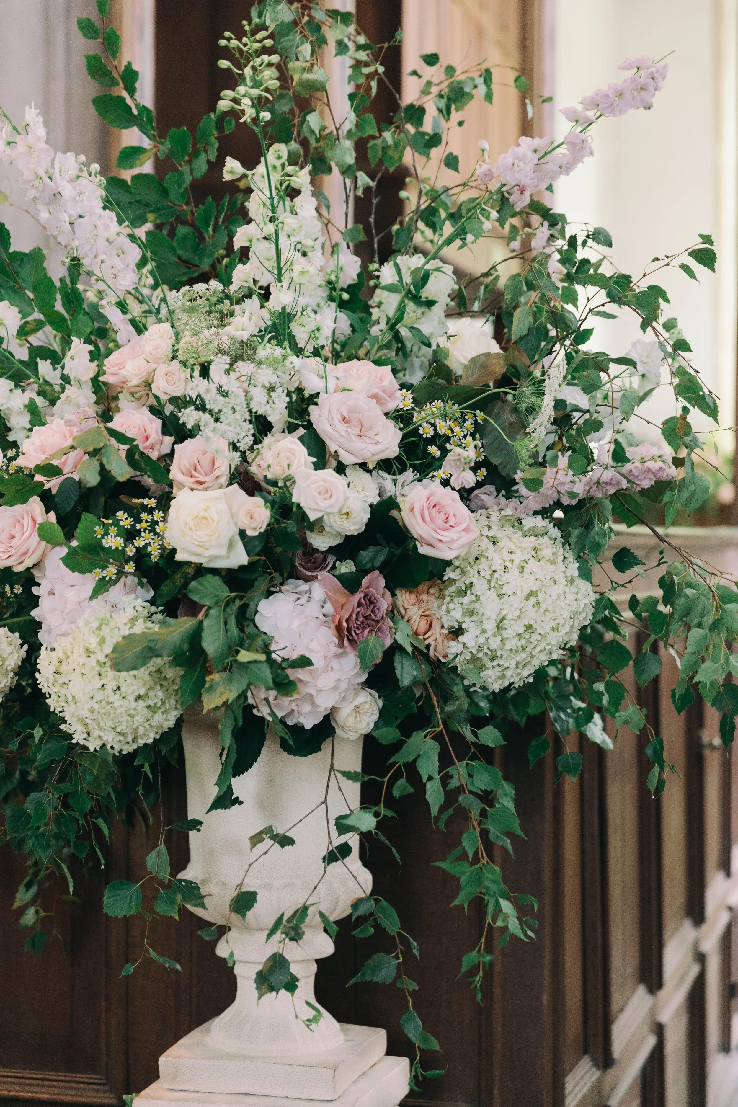 A large urn arrangement of white and pale pink roses, hydrangeas, and various greenery in a white vase, set in a village church in the norfolk countryside