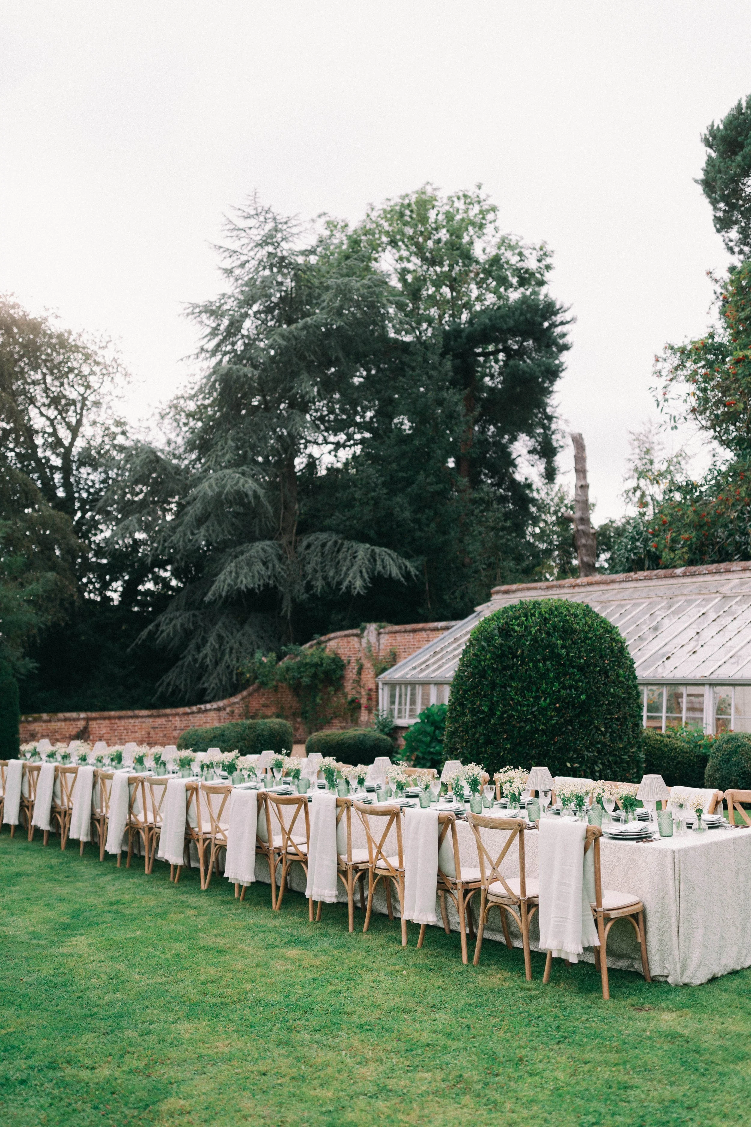Long outdoor dining table set up in a garden with floral tablecloths, floral centerpieces, and white lampshades, surrounded by greenery and trees. Hosted in Norfolk and planned by Attabara