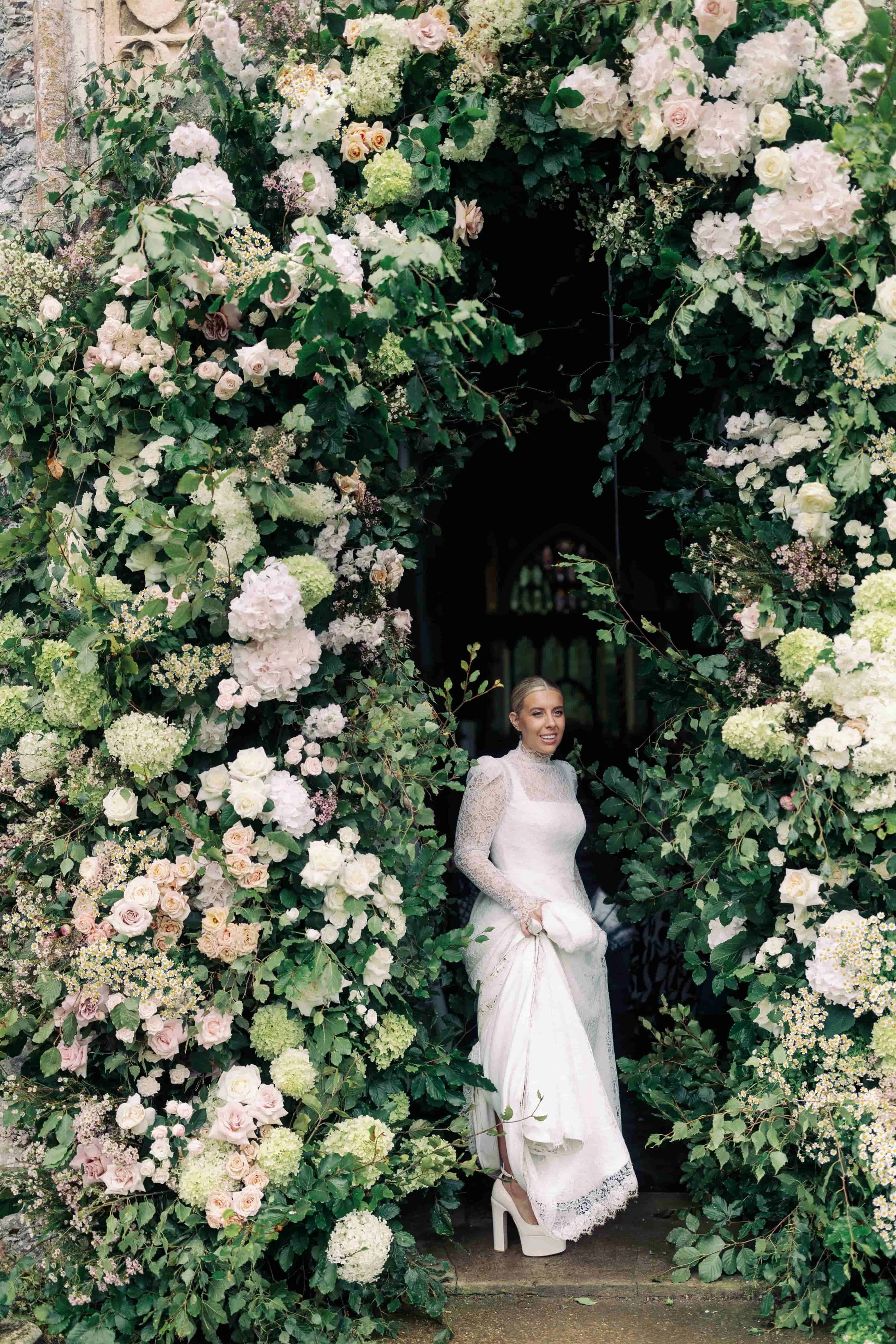 A bride in a white wedding dress standing in a floral archway composed of white and pale pink flowers and greenery. Norfolk village wedding by The Botany House Wedding Florist. Luxury wedding florals