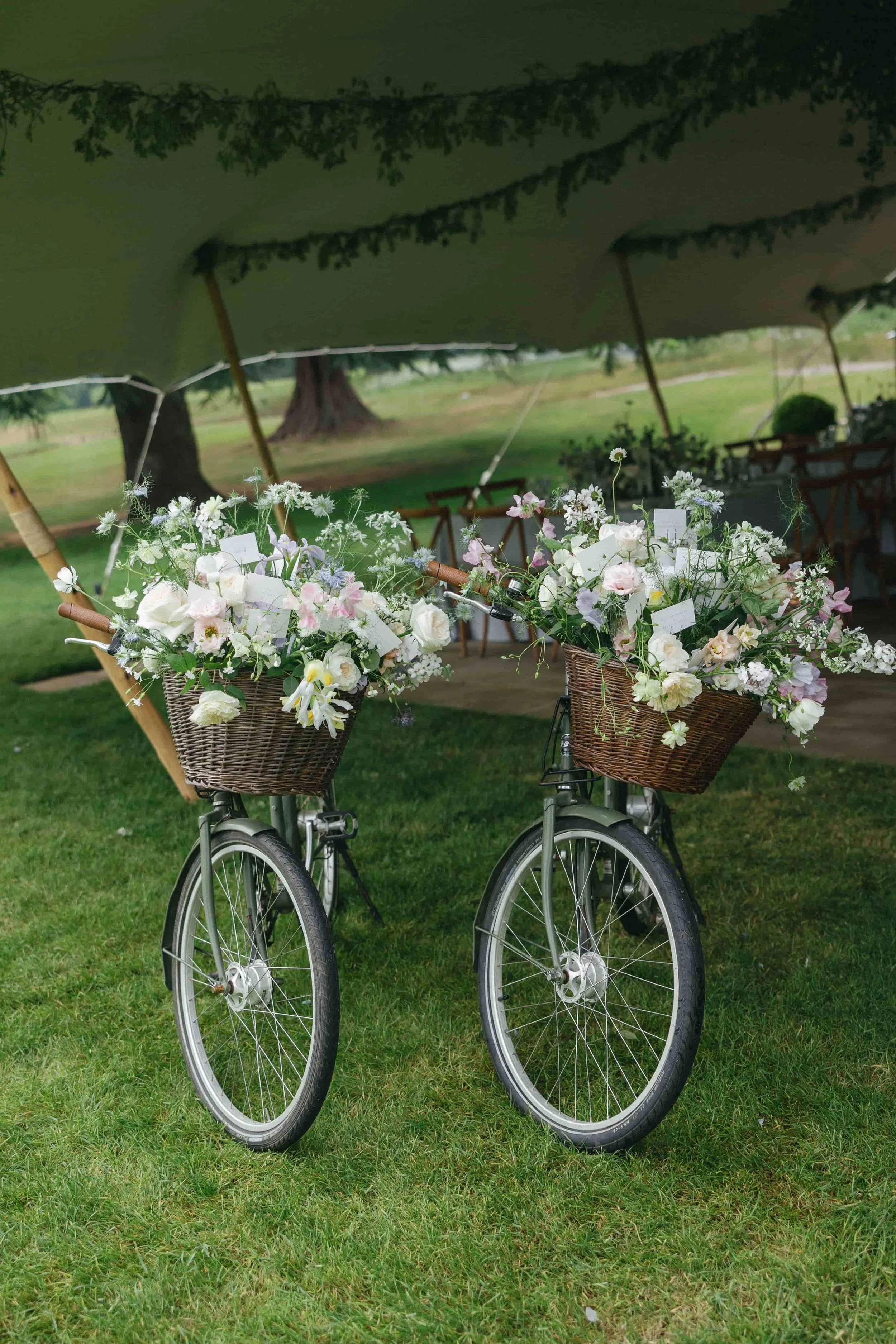 Two bicycles decorated with white and pastel-colored flowers in baskets, parked underneath a large green tent on a grassy field. Wilderness Reserve, Suffolk Florals By The Botany House