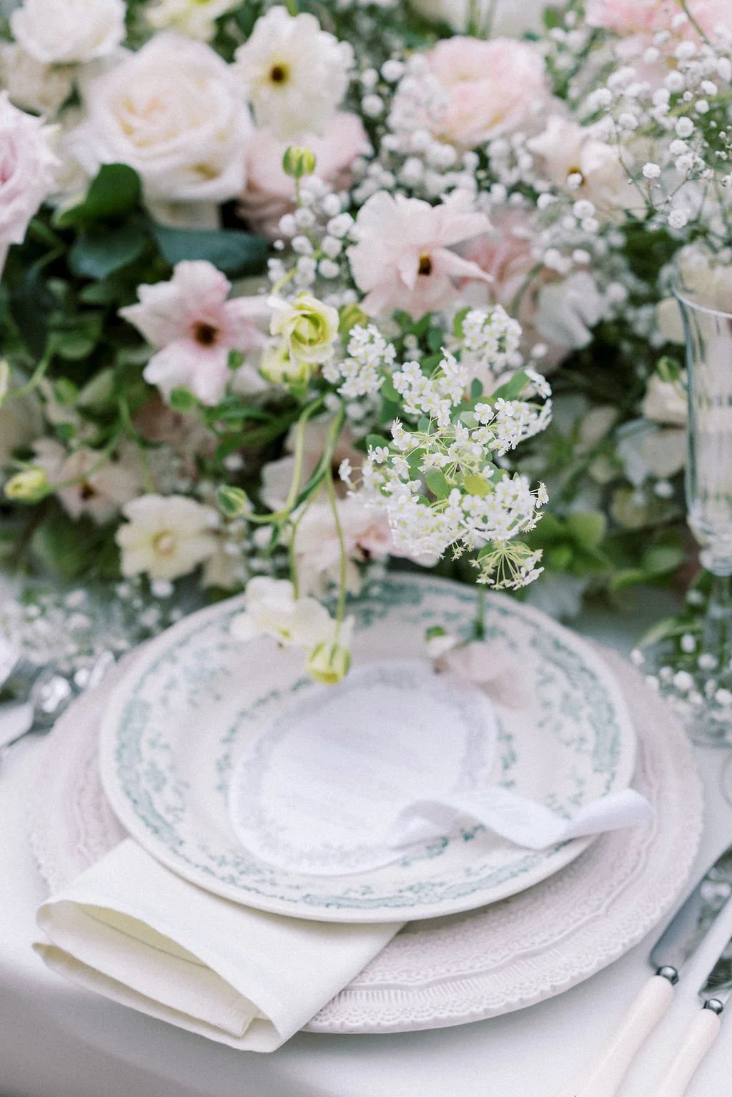 Elegant table setting with a floral centerpiece featuring white and light pink flowers, a white plate with intricate blue patterns, a folded white napkin, and silverware, all on a white tablecloth.