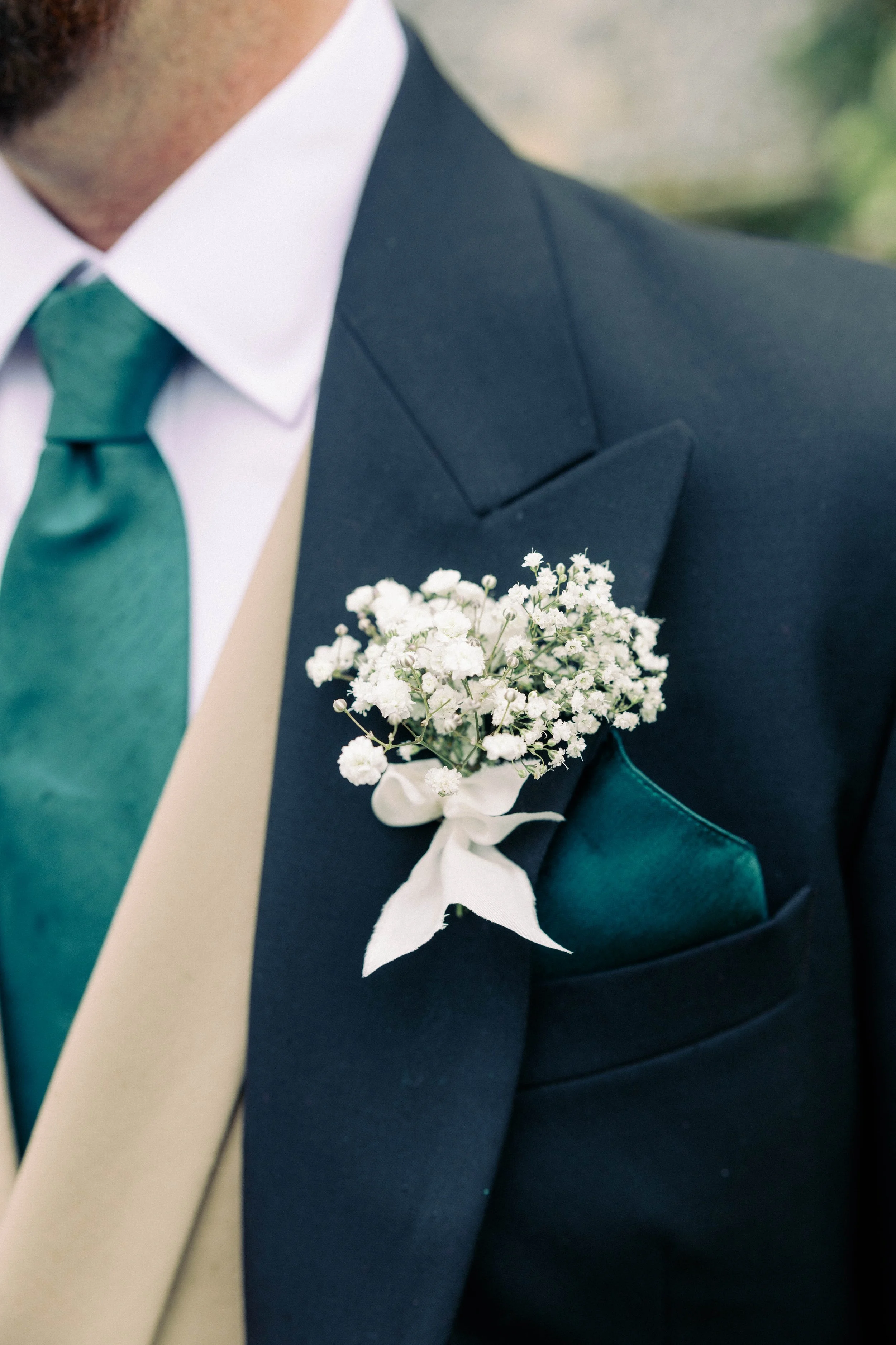 Close-up of a person's dark blue suit with a teal pocket square, white shirt, teal tie, and a white buttonhole with small white flowers and a white ribbon on the lapel.