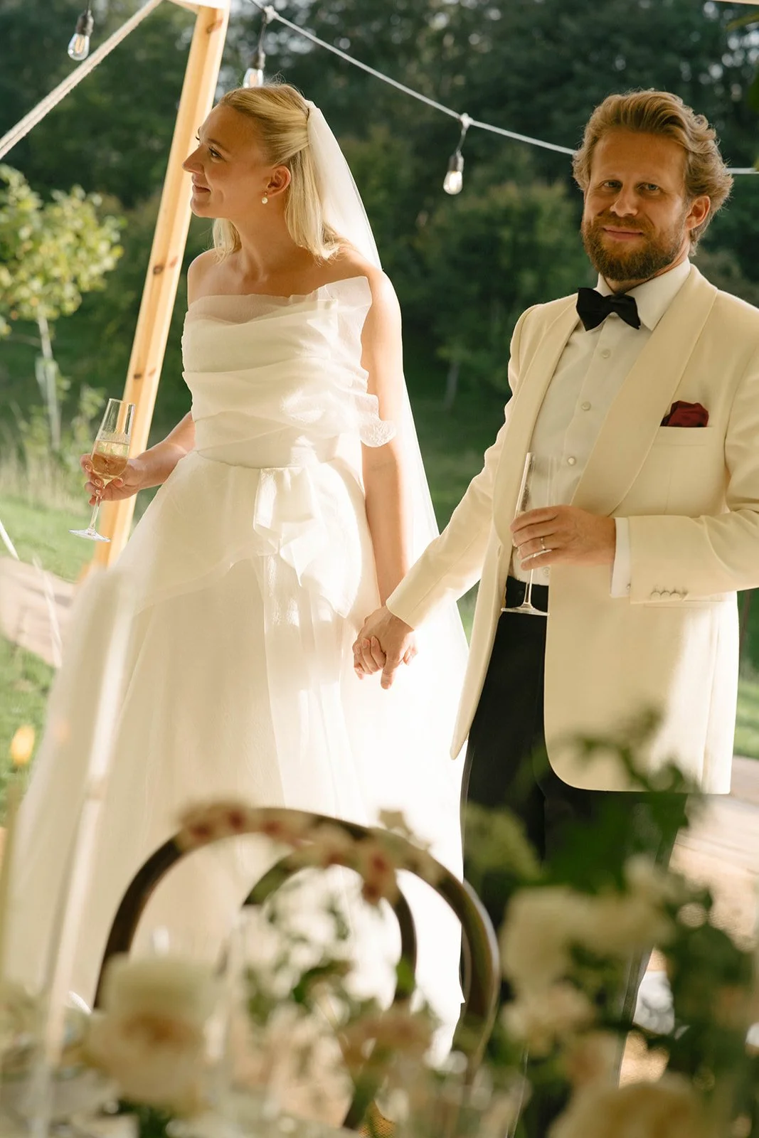 A newlywed couple holding hands during their outdoor wedding ceremony. The bride is wearing a white wedding dress and veil, holding a glass of champagne, and smiling to her left. The groom is dressed in a white tuxedo jacket with a black bow tie, hol
