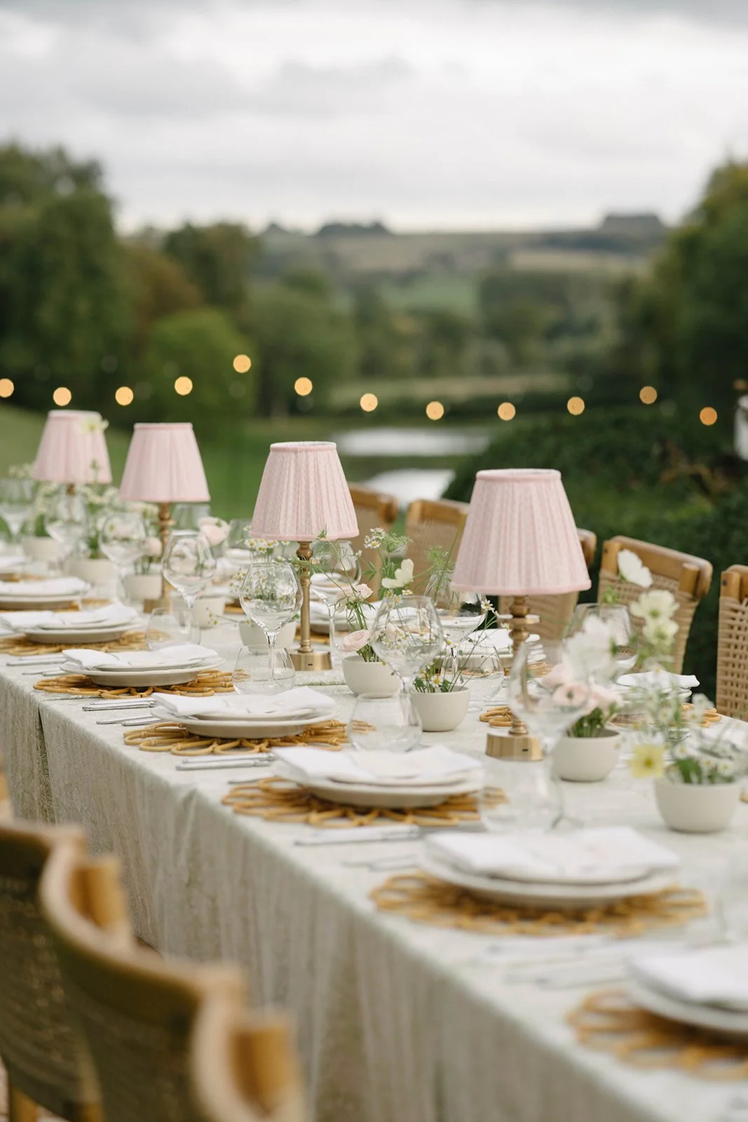 Outdoor banquet table set with white plates, glasses, cutlery, pink lamps, and floral centerpieces, overlooking a green landscape - luxury garden-style wedding flowers by The Botany House, wedding florist for Cornwell Manor, Oxfordshire