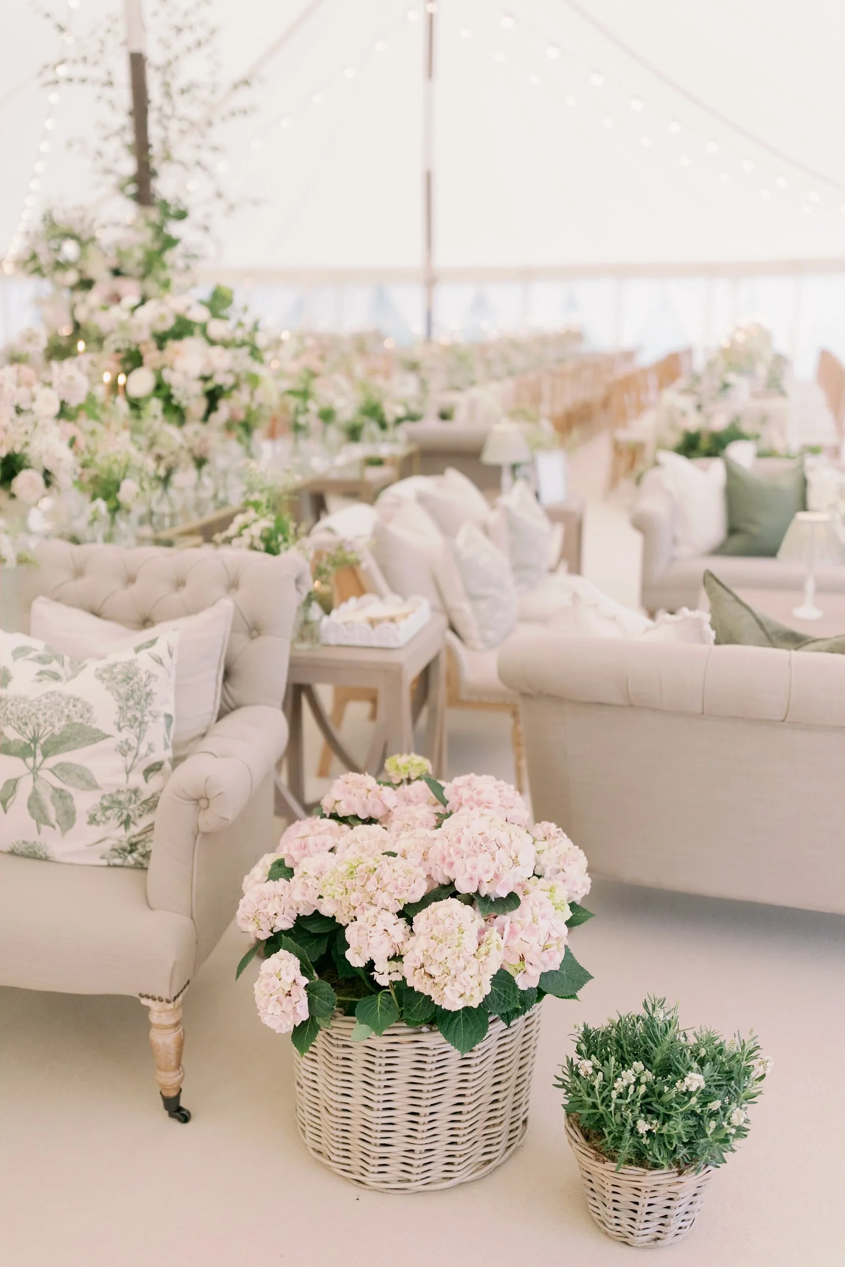 Elegant, light-colored sofas with floral pillows and wicker baskets of pink hydrangeas in a bright, sailcloth marquee. Hosted in Norfolk