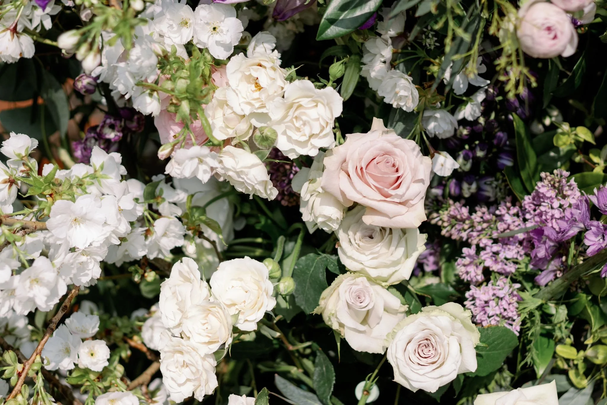 A variety of white, pink, and purple flowers including roses and small blossoms, with green leaves.