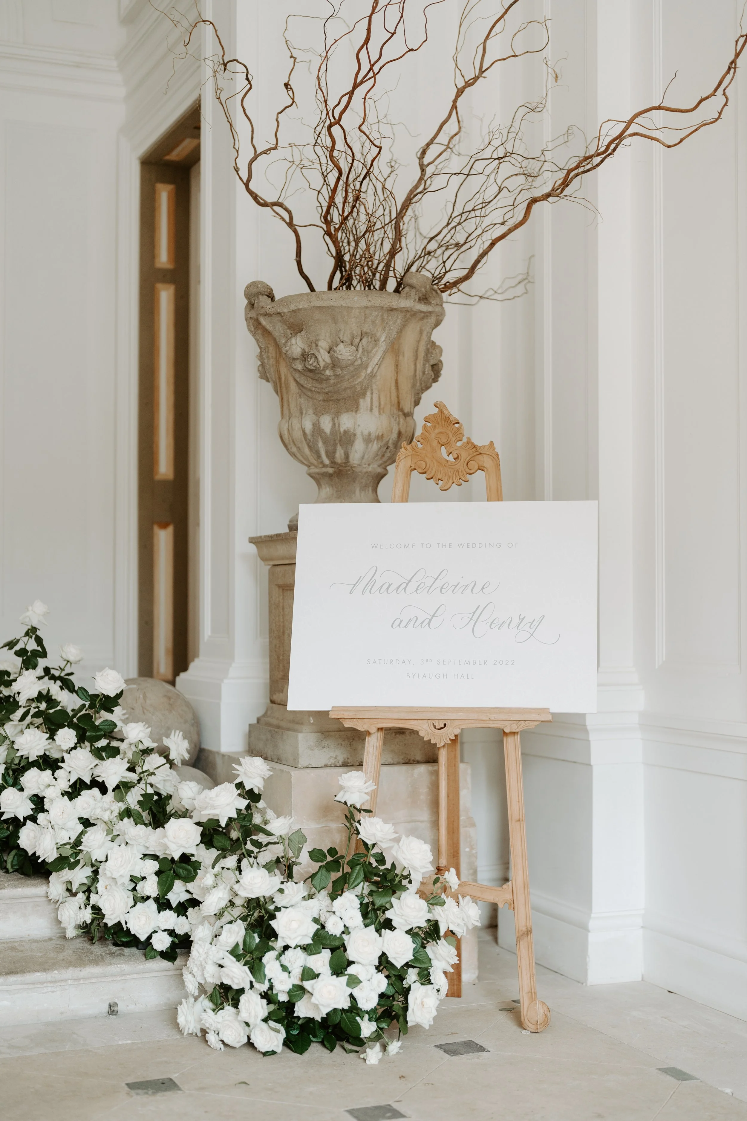 Wedding welcome sign on an easel with white flowers and decorative vase with branches in a white, elegant room.