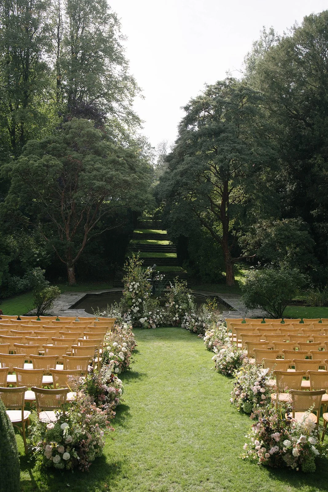 Outdoor wedding ceremony setup at Cornwell Manor with rows of chairs and luxury garden-style floral arrangements along a grassy aisle, framed by trees and a small fountain, by The Botany House wedding florist.