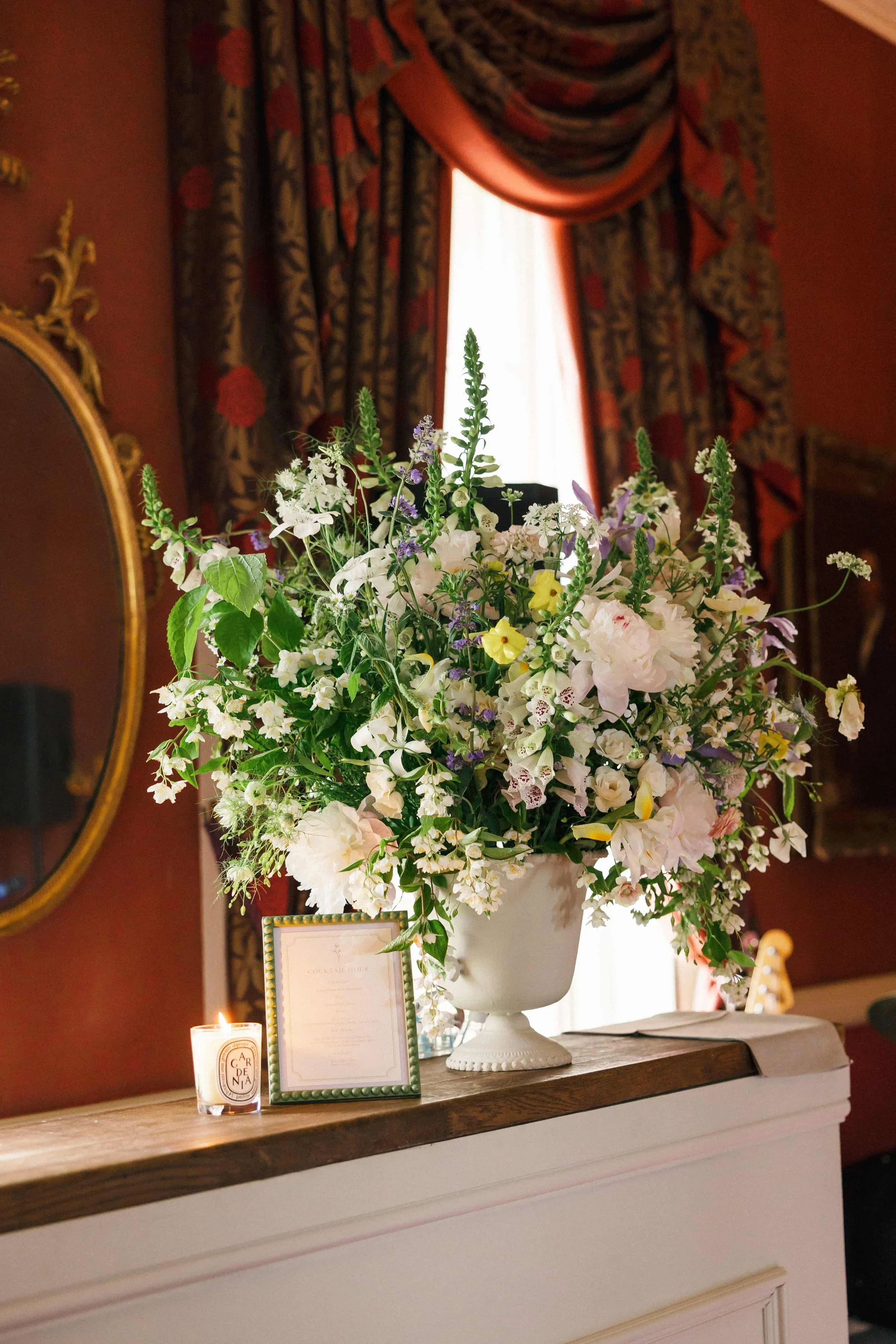 A large floral arrangement in a white vase on a mantle, with a lit candle and framed menu or note, in a room with ornate red and gold curtains and mirror.