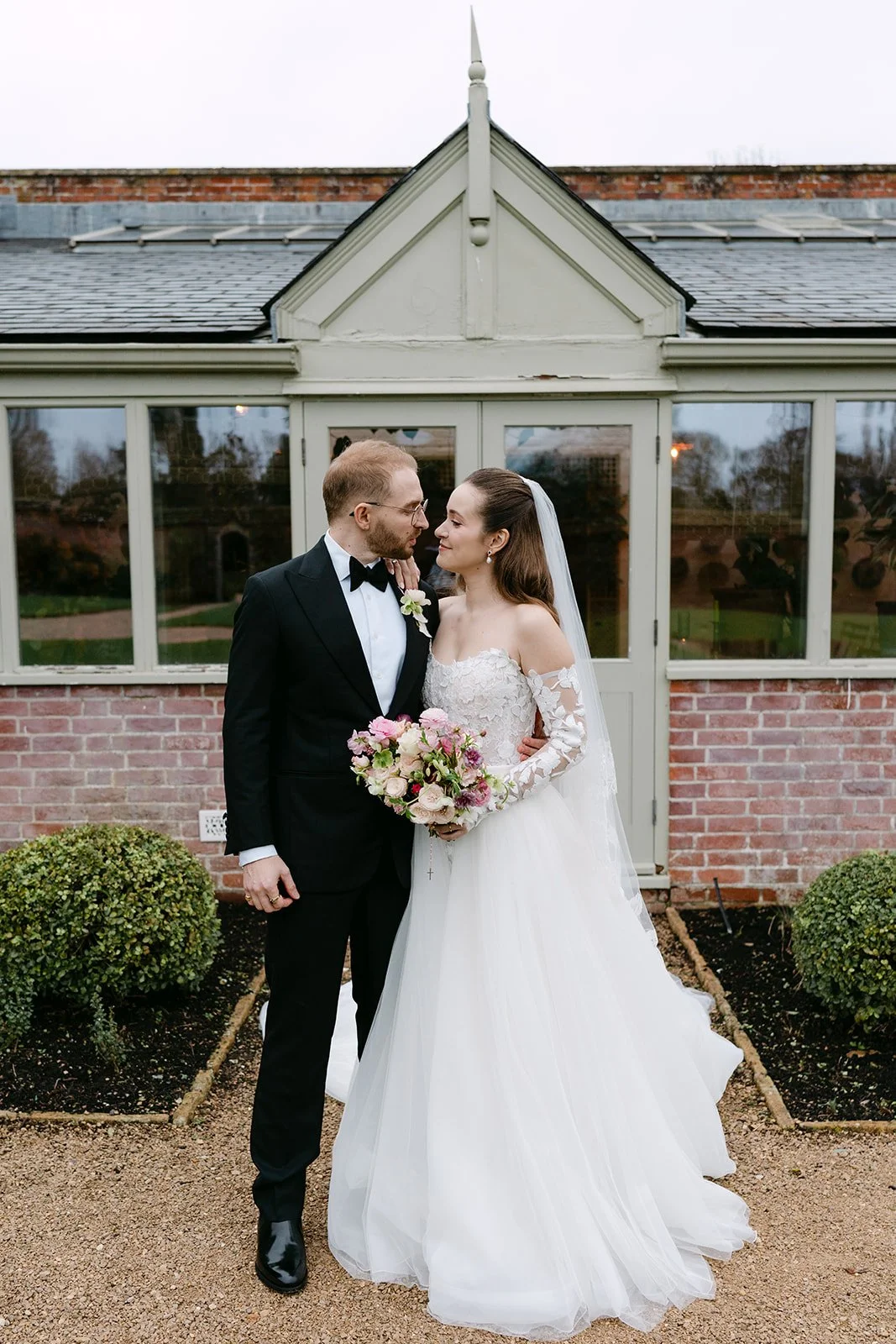 A bride and groom stand close together in wedding attire, looking into each other's eyes, with the bride holding a bouquet of pink and white flowers. They are outdoors in front of a brick building with large windows. Kin House, The Botany House