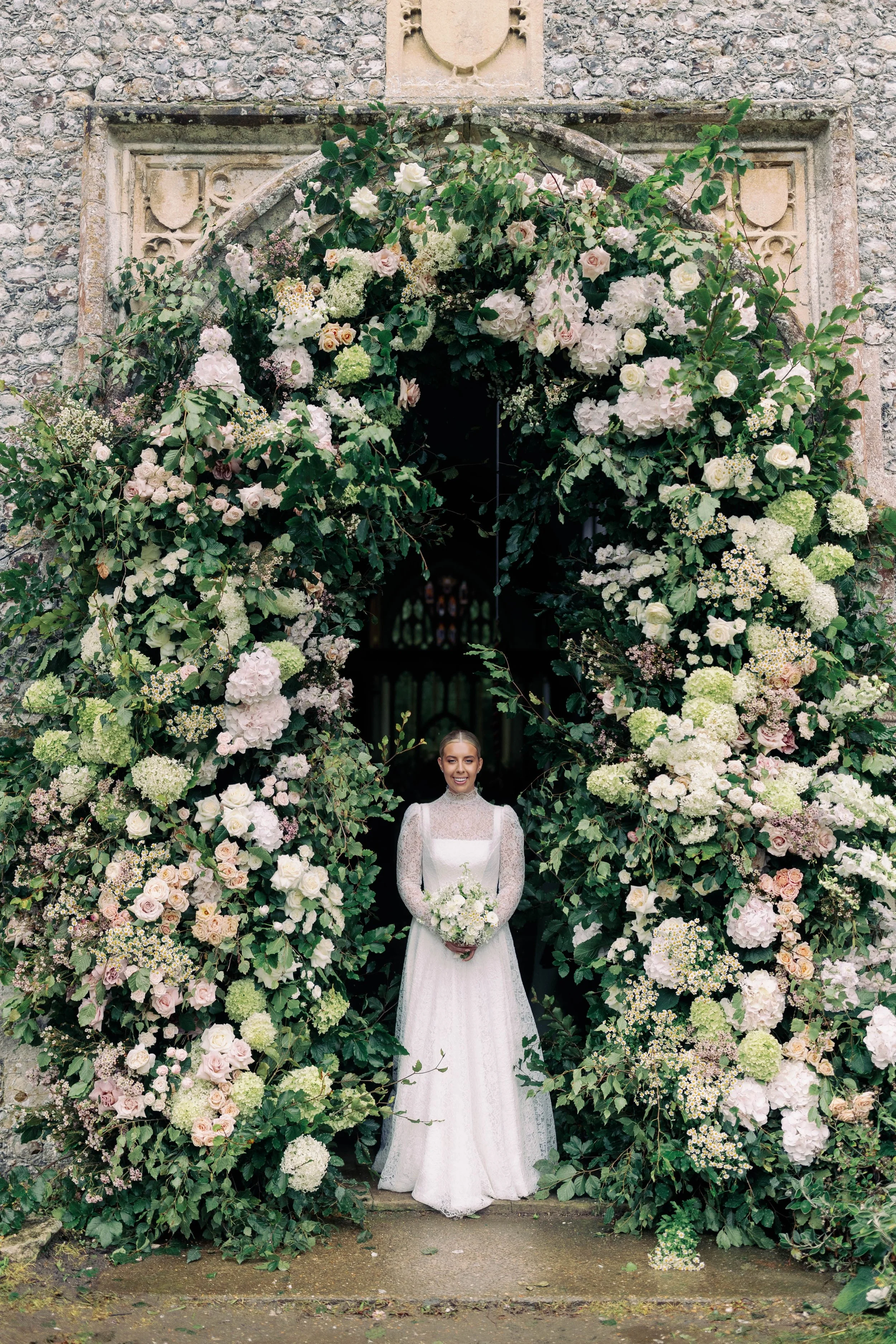 Bride in a white wedding dress holding a bouquet standing under a large floral archway at a church entrance in Heydon Norfolk