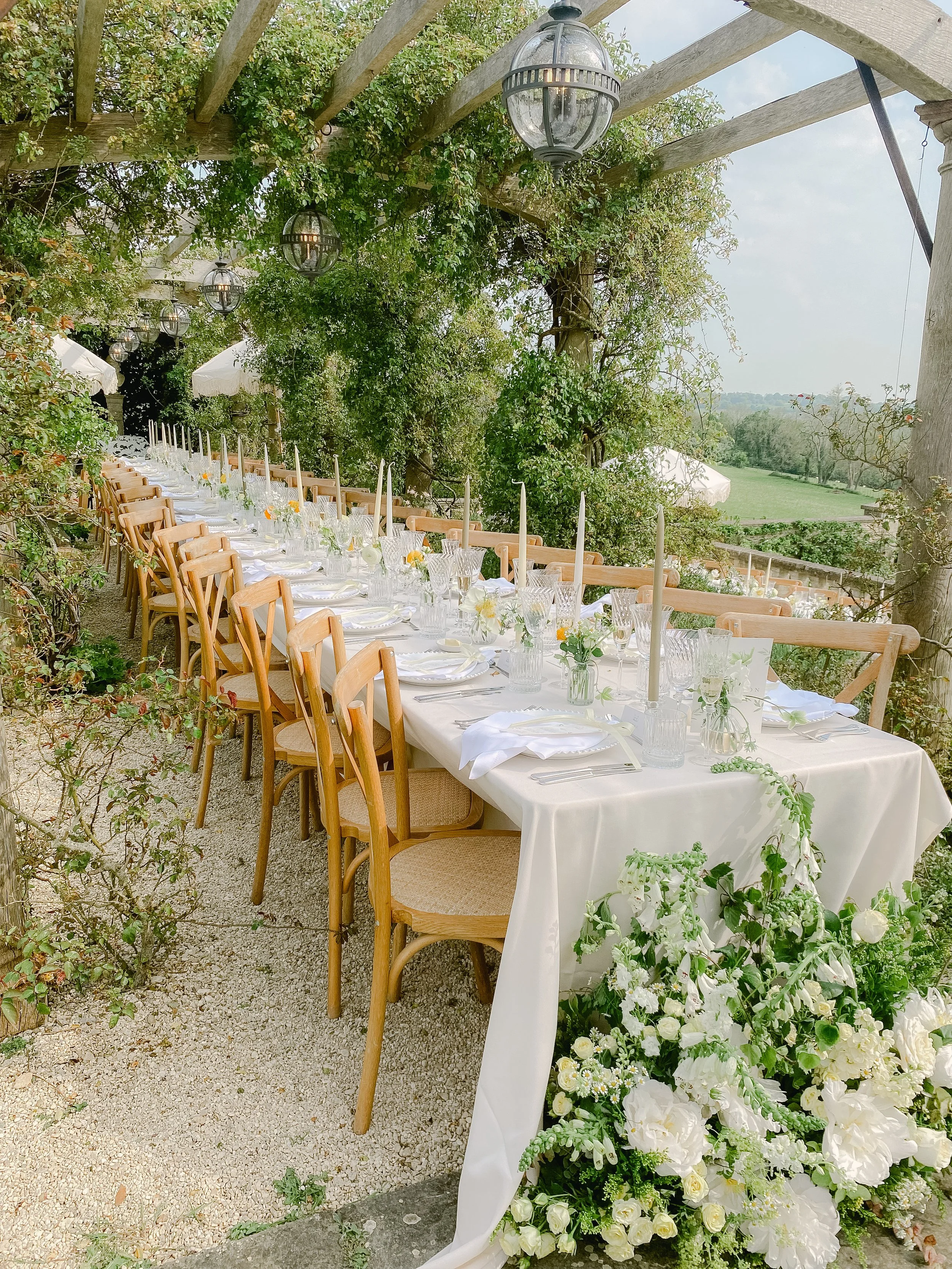 An outdoor dining setup with a long white tablecloth-covered table decorated with white flowers and greenery, set under a wooden pergola with hanging lanterns, surrounded by lush greenery and open view of a field.