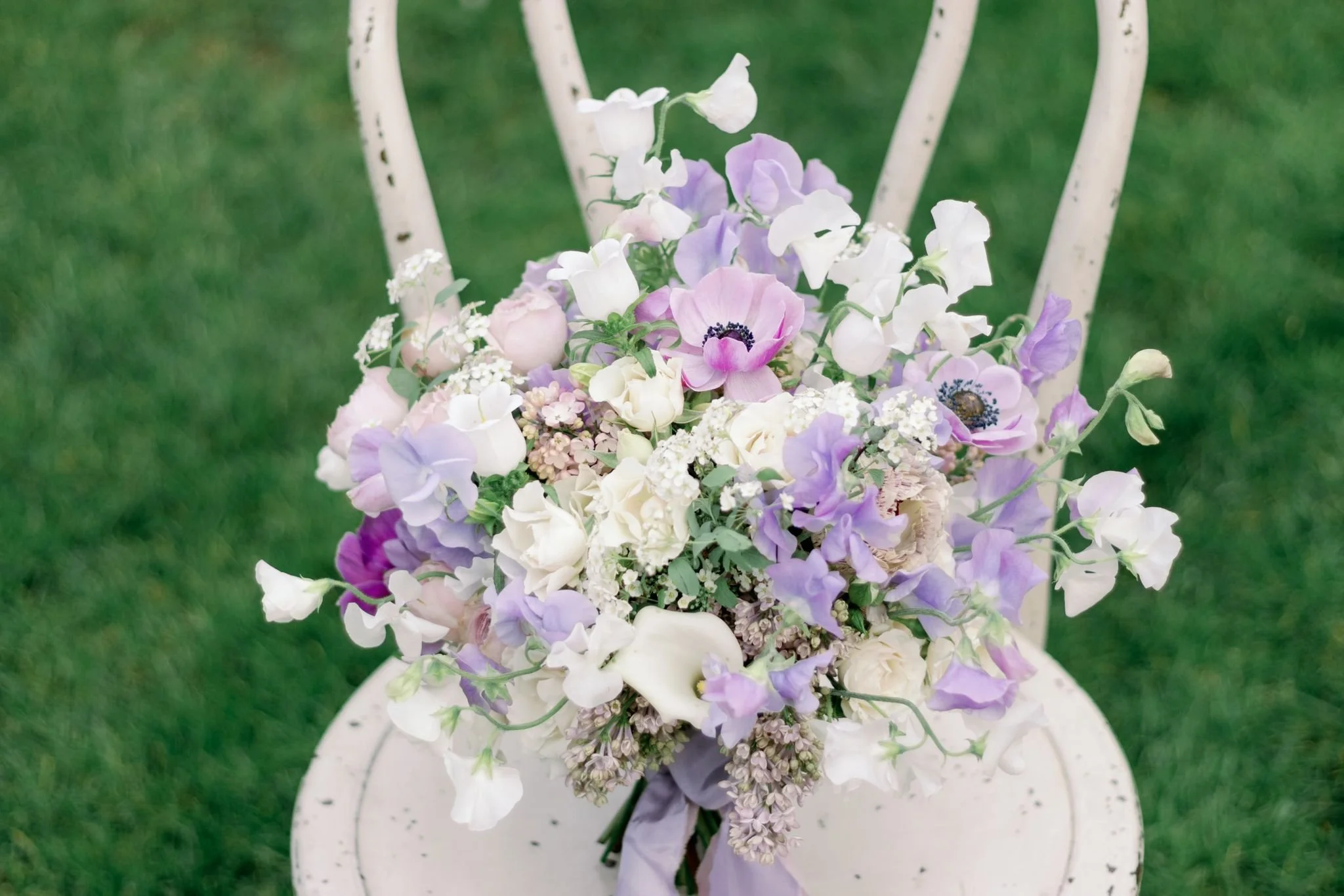A bouquet of white, lavender, and light pink flowers arranged on a white, distressed metal chair on a green lawn.
