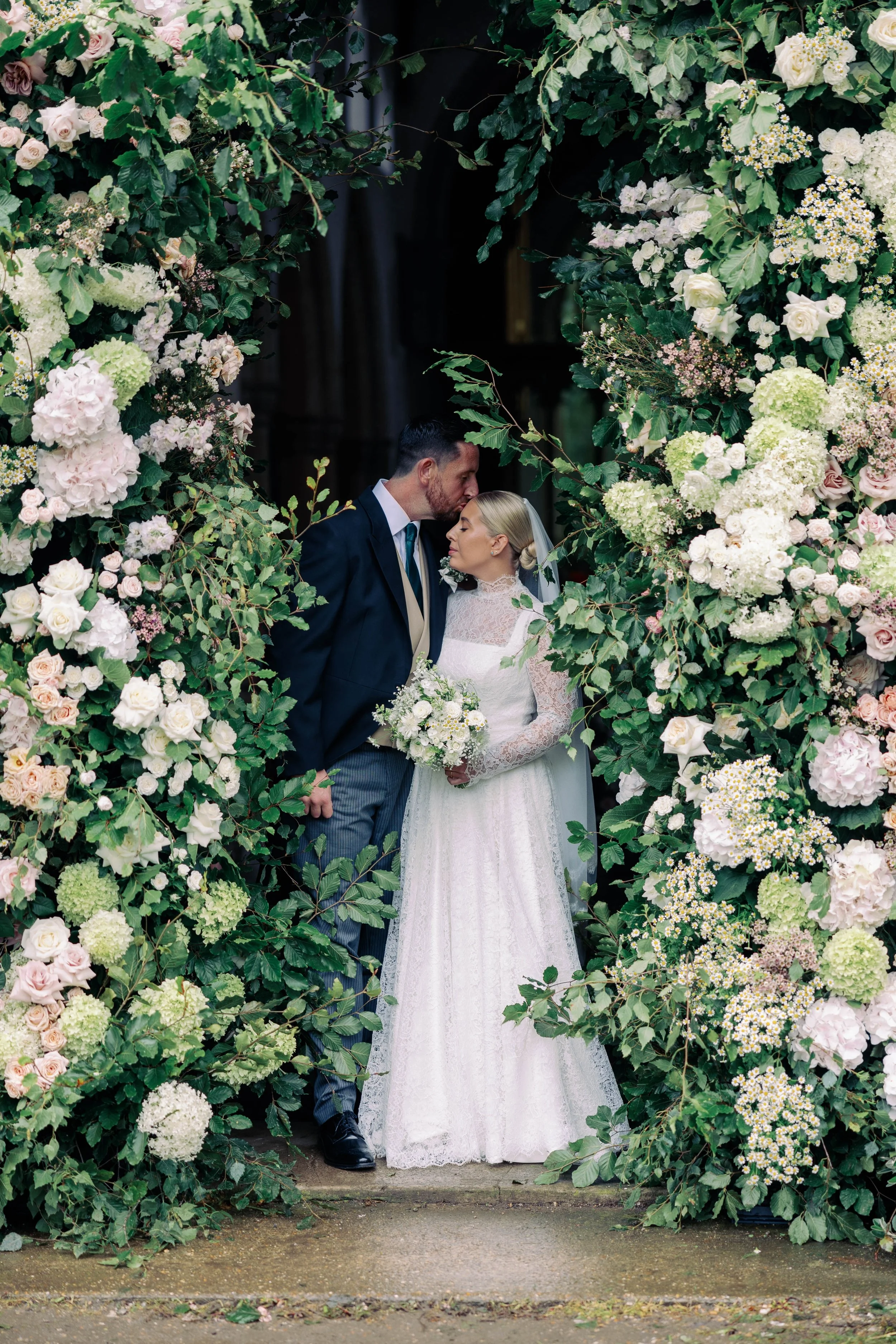 A bride in a white lace wedding dress and veil holding a bouquet standing close to a groom in a dark suit with a light-colored vest, in front of a floral archway with pink and white flowers and green leaves, during a wedding ceremony.