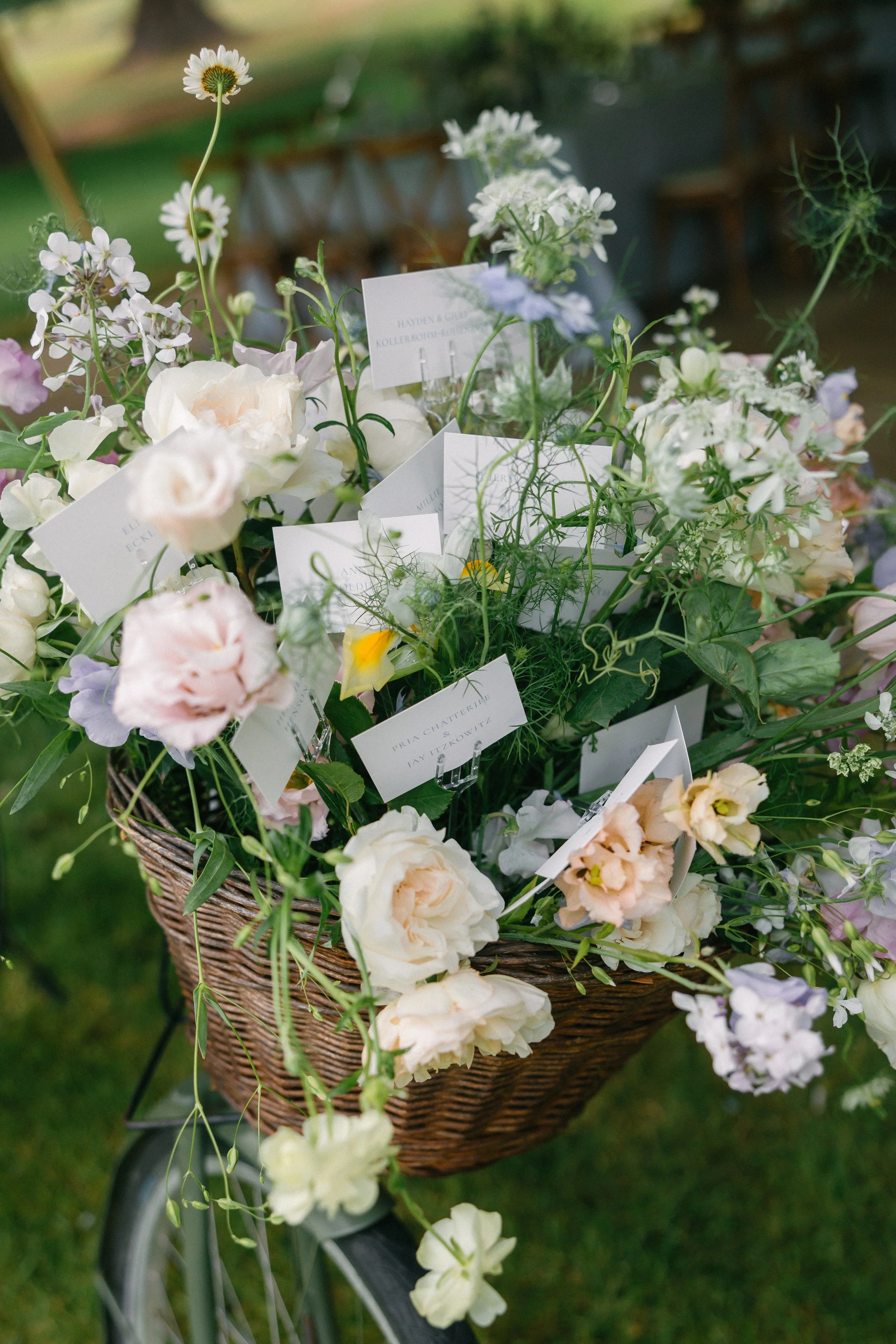 A bicycle basket filled with a variety of white, blush, and pastel-colored flowers, with small white cards inserted among the blooms.