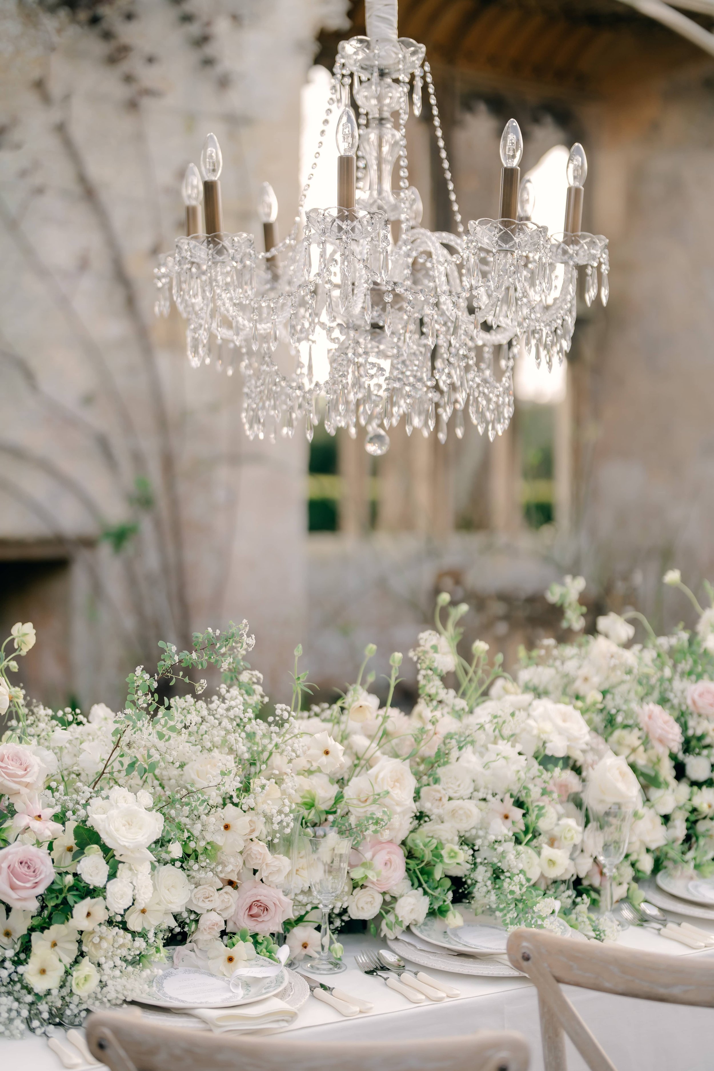 Elegant table setting with a large floral arrangement of white and pale pink flowers, a crystal chandelier hanging above, and a rustic stone wall in the background. Sudeley Castle The Cotswolds, Florals by The Botany House