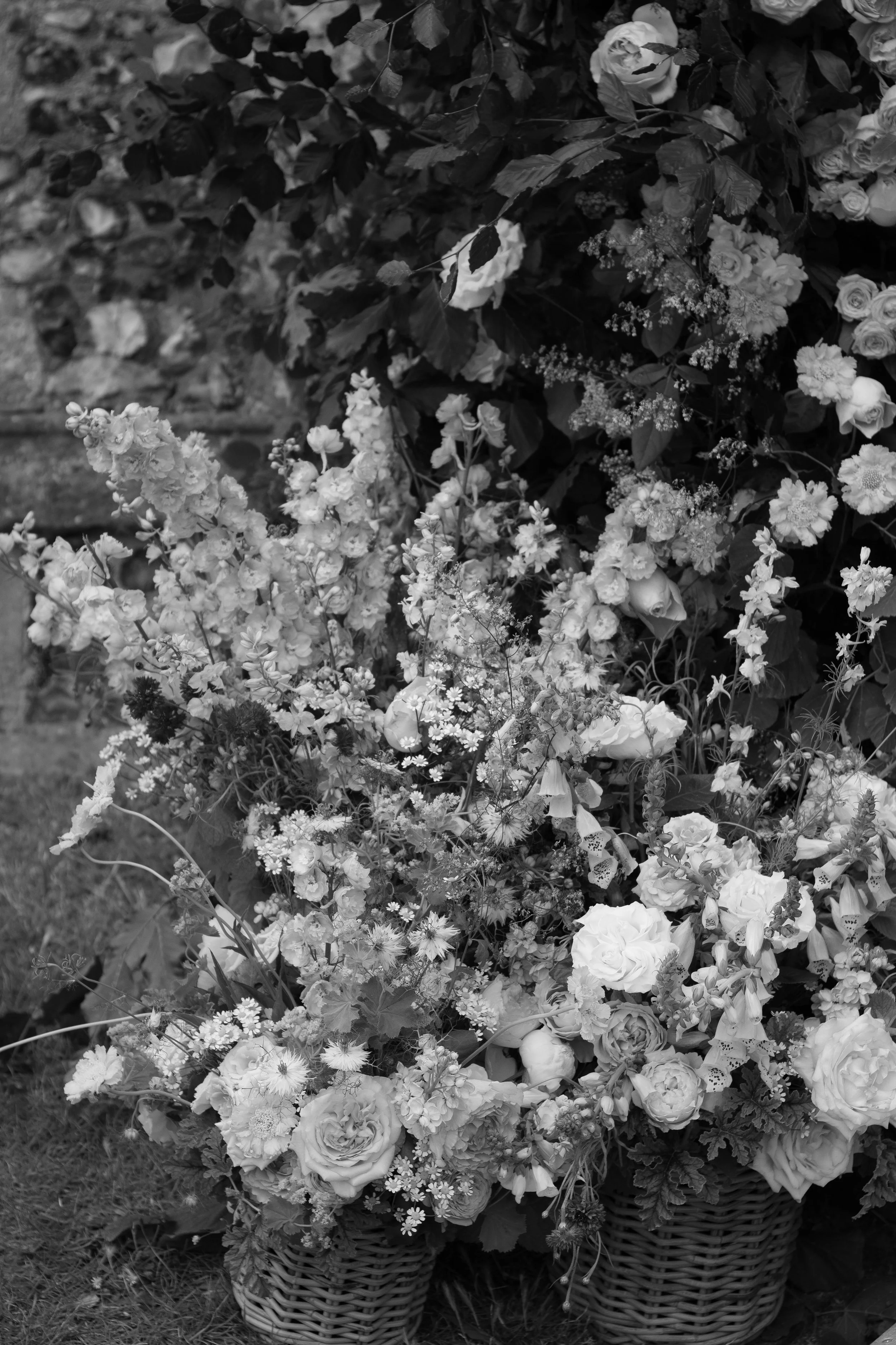 A large arrangement of various flowers in baskets, with a mix of roses, daisies, and other blooms, against a background of dark leaves and foliage.