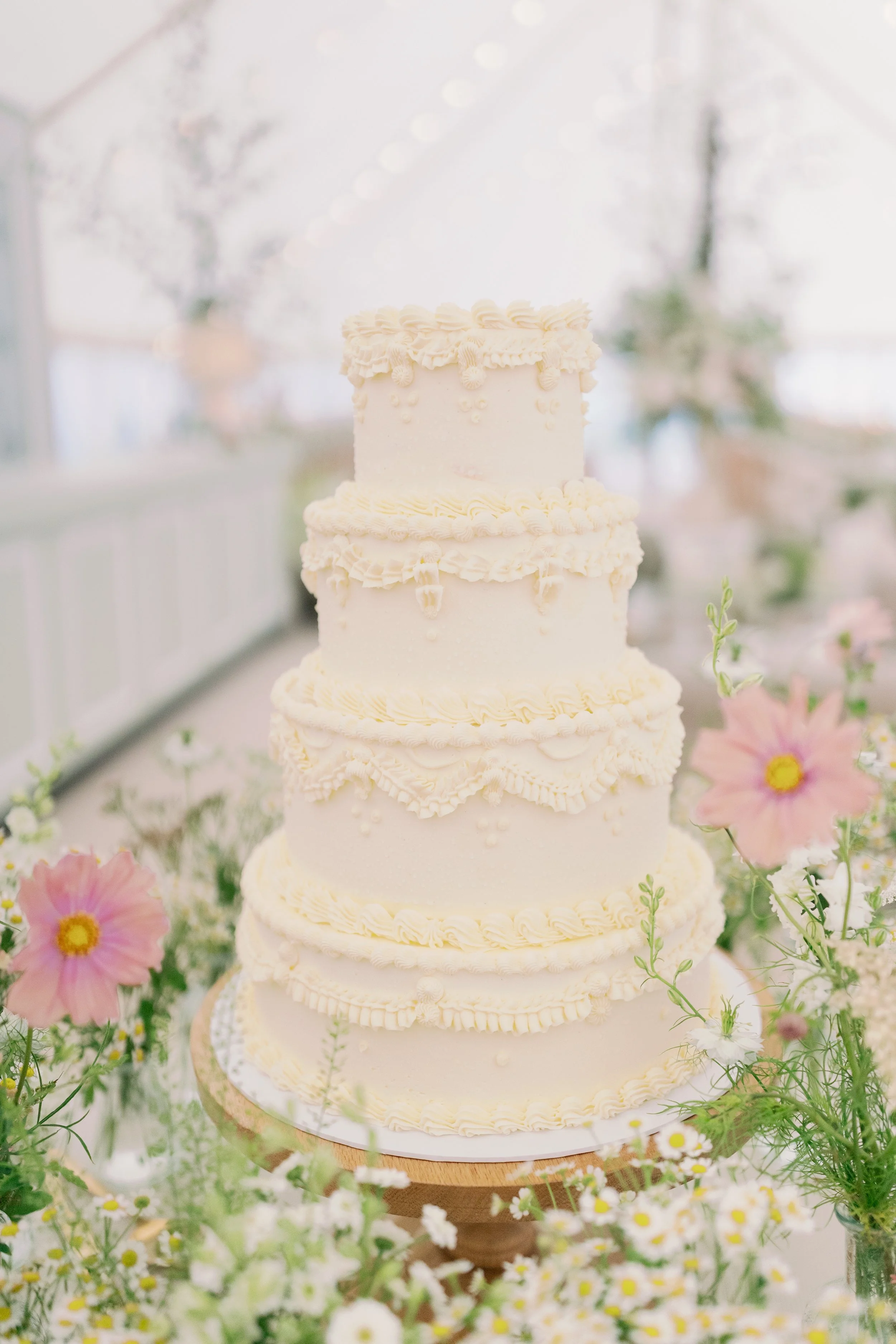 A tall, elegant, four-tiered white wedding cake with intricate piping details, surrounded by pink and white flowers on a table. Hosted in Norfolk
