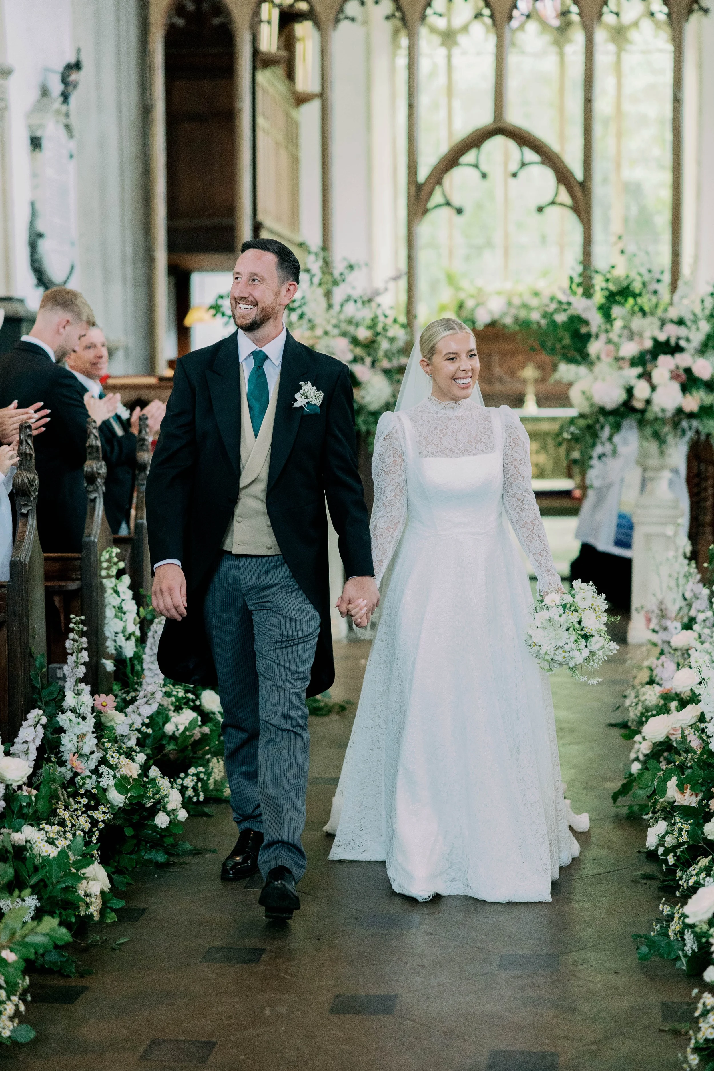 Bride and groom holding hands walking down the aisle in a wedding ceremony, surrounded by floral arrangements and guests applauding.