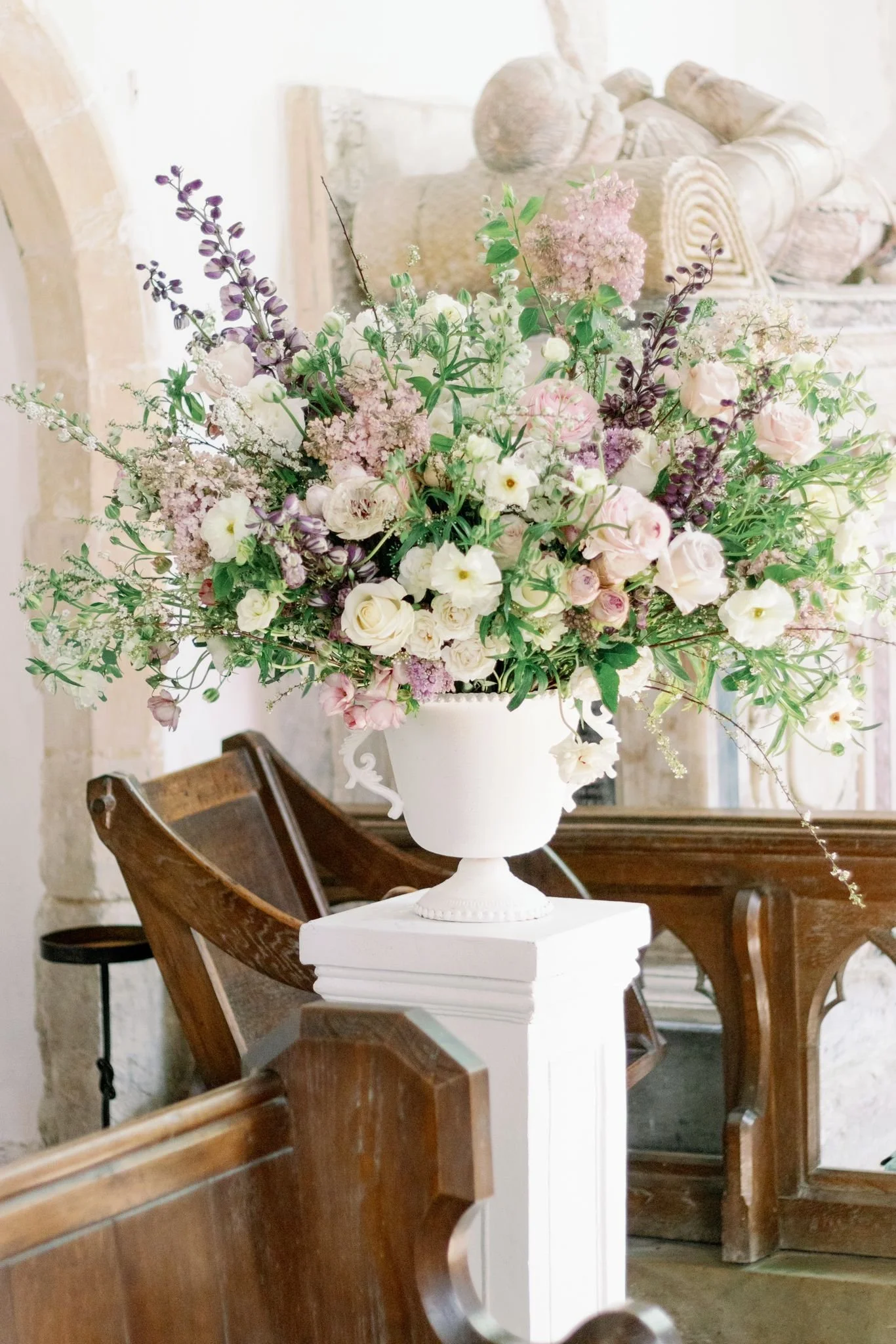 A large bouquet of mixed flowers in shades of white, pink, purple, and green in a white urn on a white pedestal, inside a rustic room with wooden furniture and stone walls.