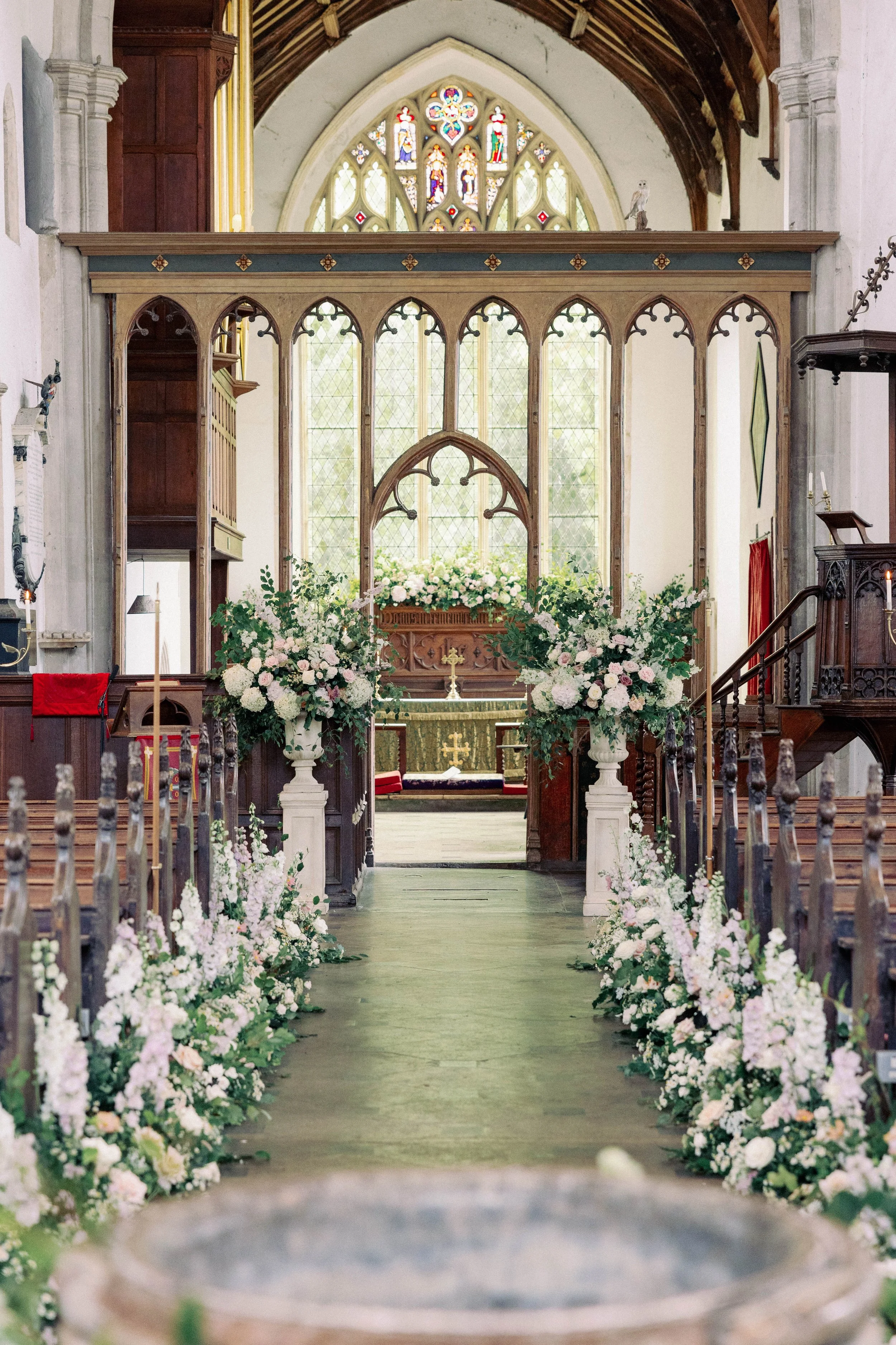 Inside a historic church decorated for a wedding, with meadows lining the aisle and an altar at the front illuminated by stained glass windows. Large Urn arrangements at the front of the church