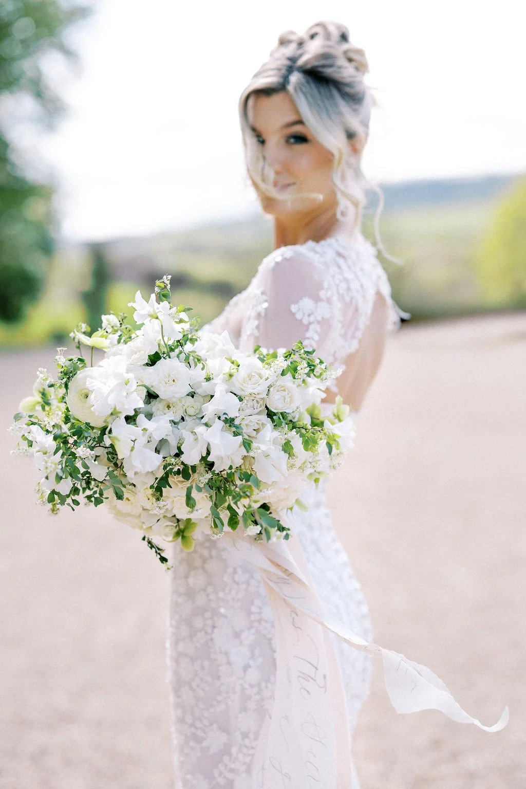 Bride in a lace wedding dress holding a large bouquet of white flowers outdoors.