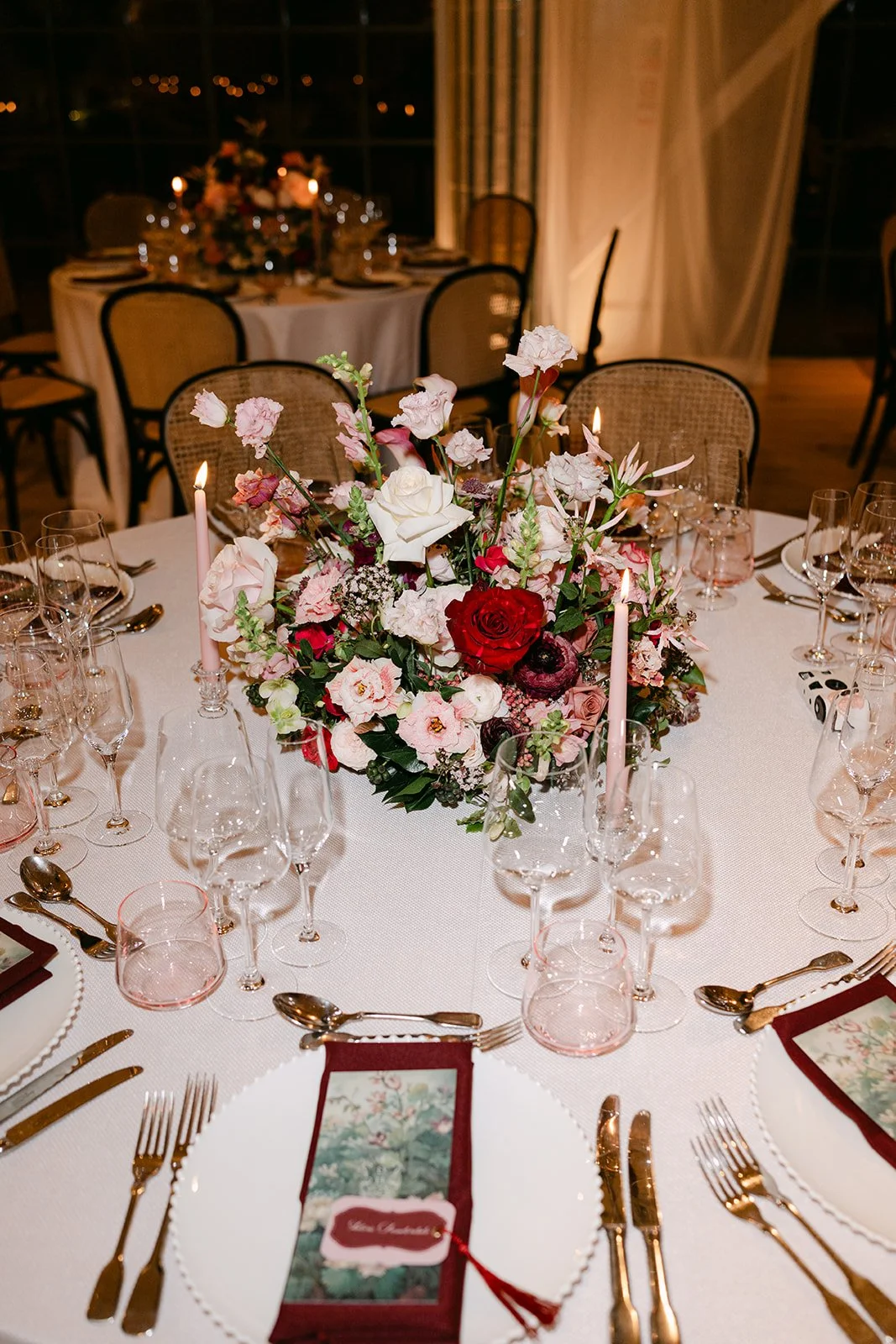 A round dining table decorated with a floral centerpiece, candles, glassware, and gold cutlery, set for a formal event at night. The Botany House Wedding Florist, Kin House The Cotswolds. Planned by Get Wed