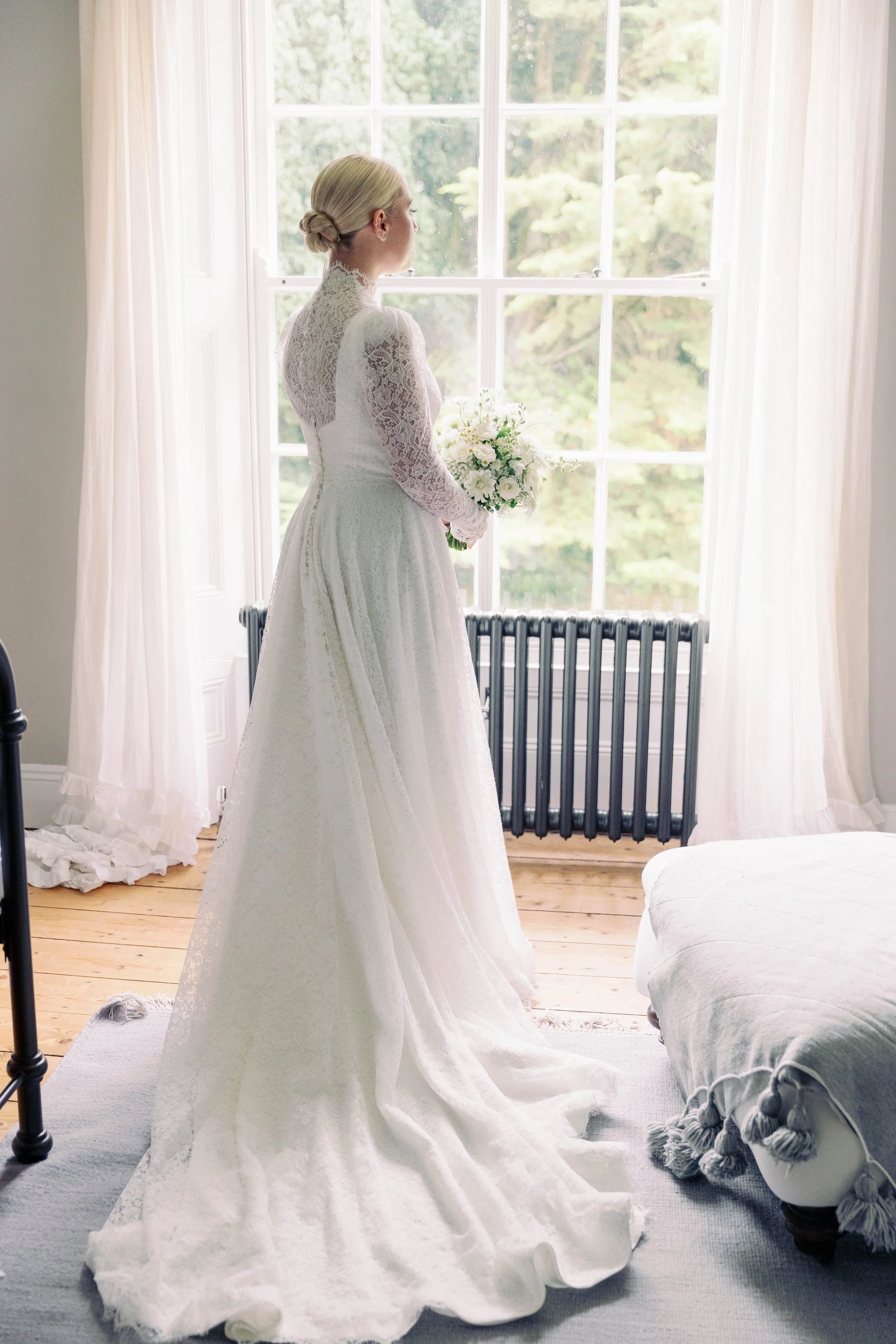 A bride in a long white lace wedding dress holding a bouquet, standing by a large window in a bright room.