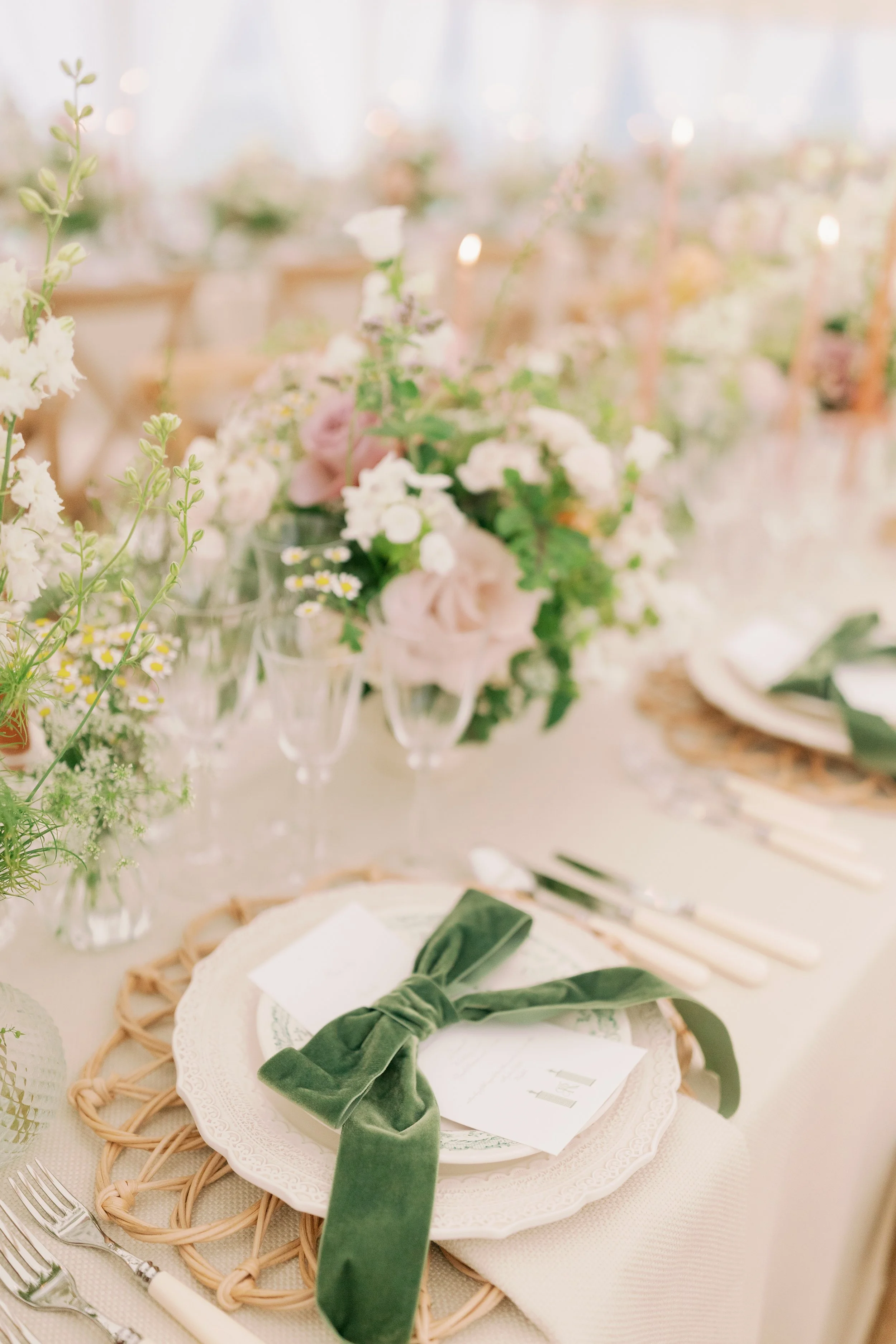Elegant table setting with floral centerpiece, green velvet bow on white plate, surrounded by glassware and cutlery, in a softly lit sailcloth marquee in Norfolk