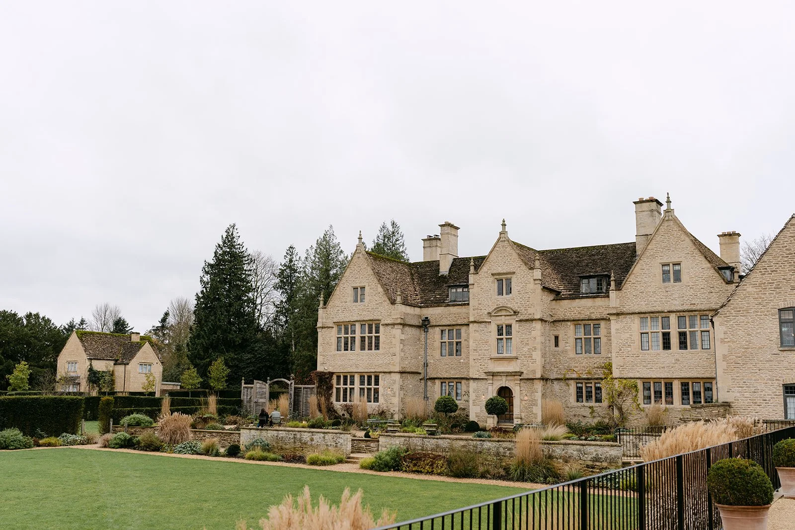 Kin House in the Cotswolds with multiple gables, chimneys, and leaded glass windows, surrounded by landscaped gardens and a lawn under an overcast sky. Florals By The Botany House, Planning by Get Wed
