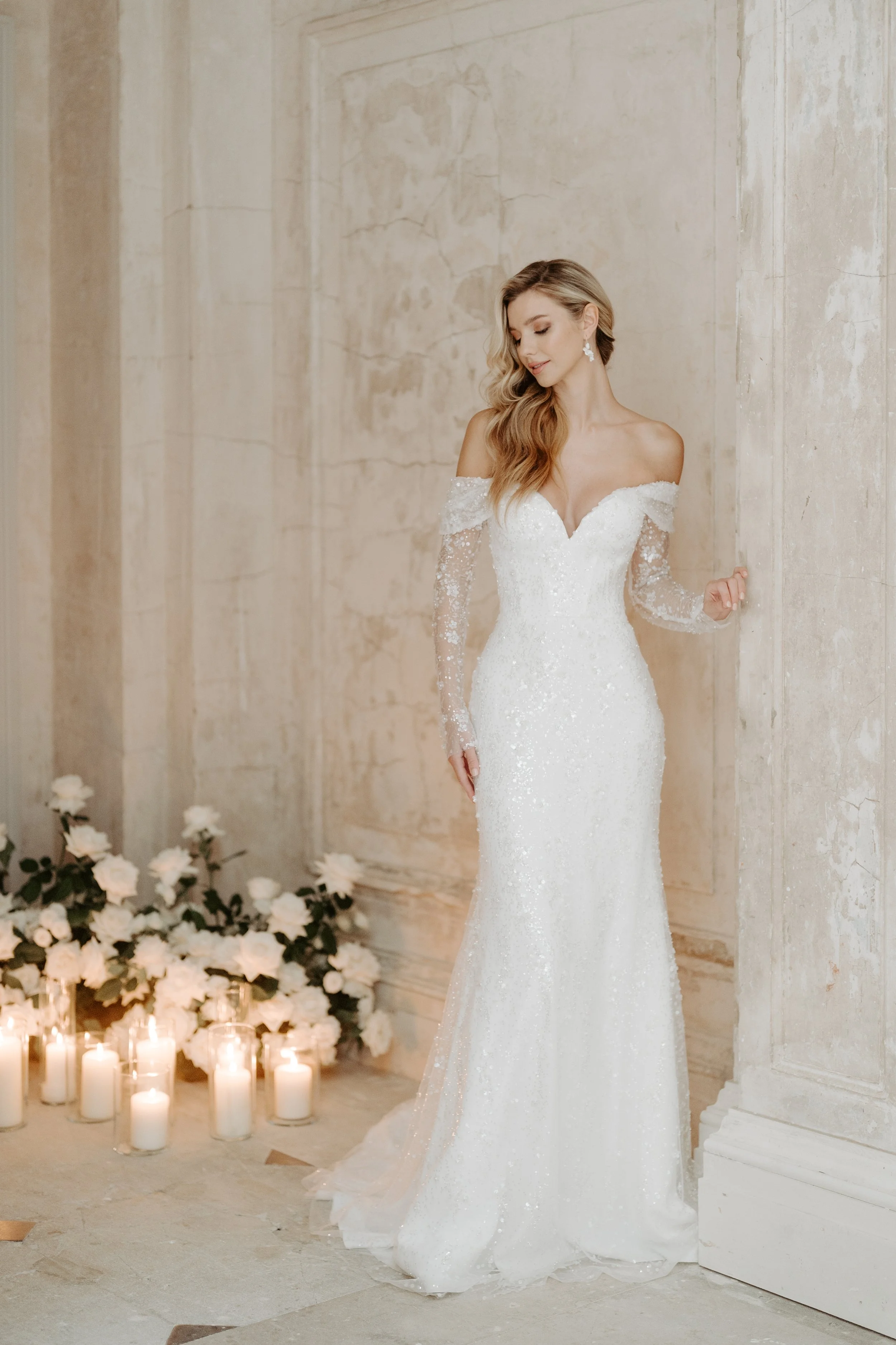A woman in a white wedding gown standing in an elegant room with candles and flowers.