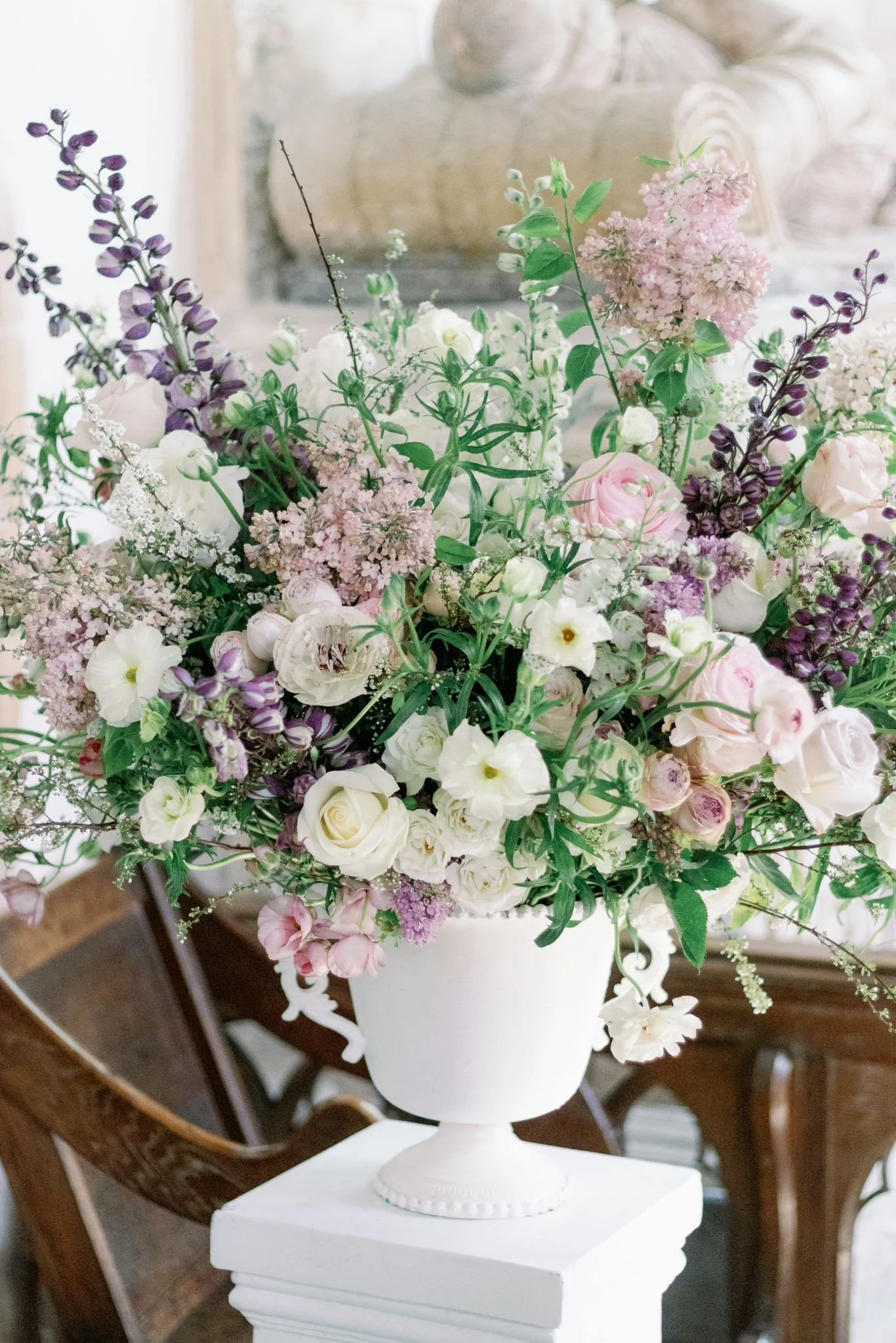 A large bouquet of mixed flowers in a white urn, including pink roses, white lisianthus, purple salvia, and other greenery, on a white pedestal in a well-lit room.