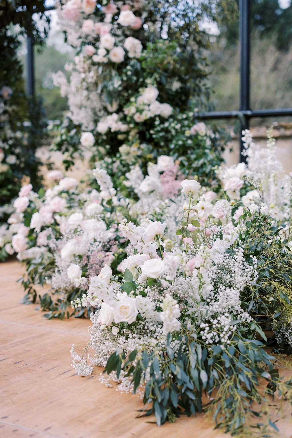 A large floral arrangement with white and pale pink flowers, along with green foliage, on a wooden surface near a window with trees outside.