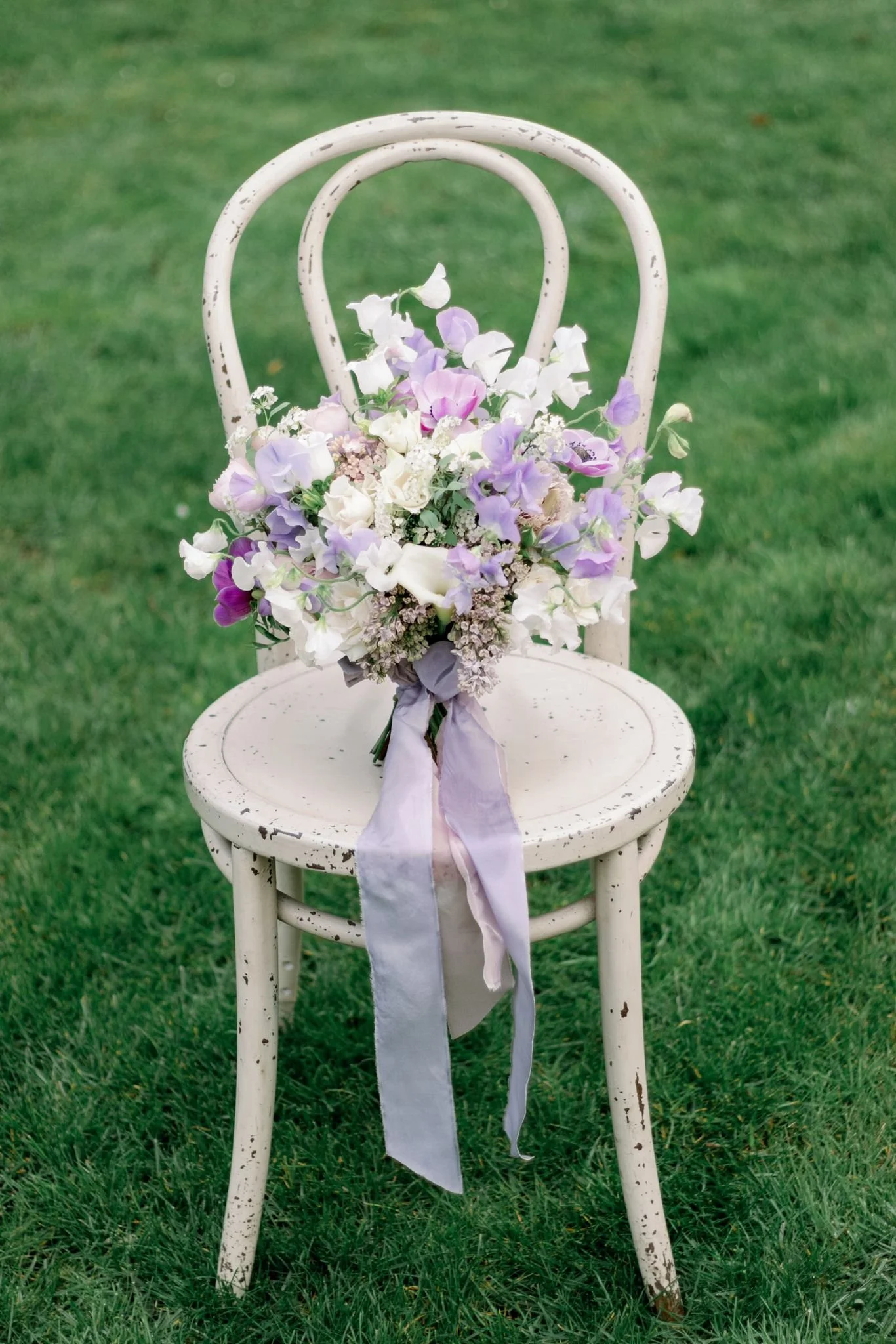 A white, distressed vintage chair with a bouquet of purple, white, and pink flowers tied with a purple ribbon resting on the seat, outdoors on grass.