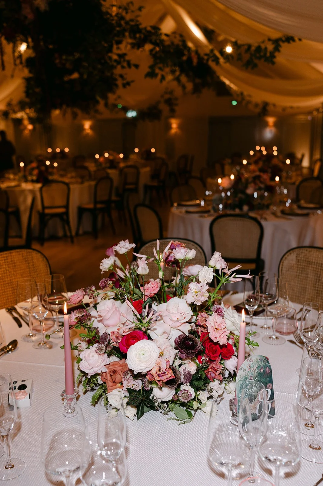 Elegant wedding reception table with pink, white, and purple flowers, pink candles, and glassware, in a decorated venue with draped ceiling and soft lighting. The Botany House Wedding Florist, Kin House The Cotswolds. Planned by Get Wed