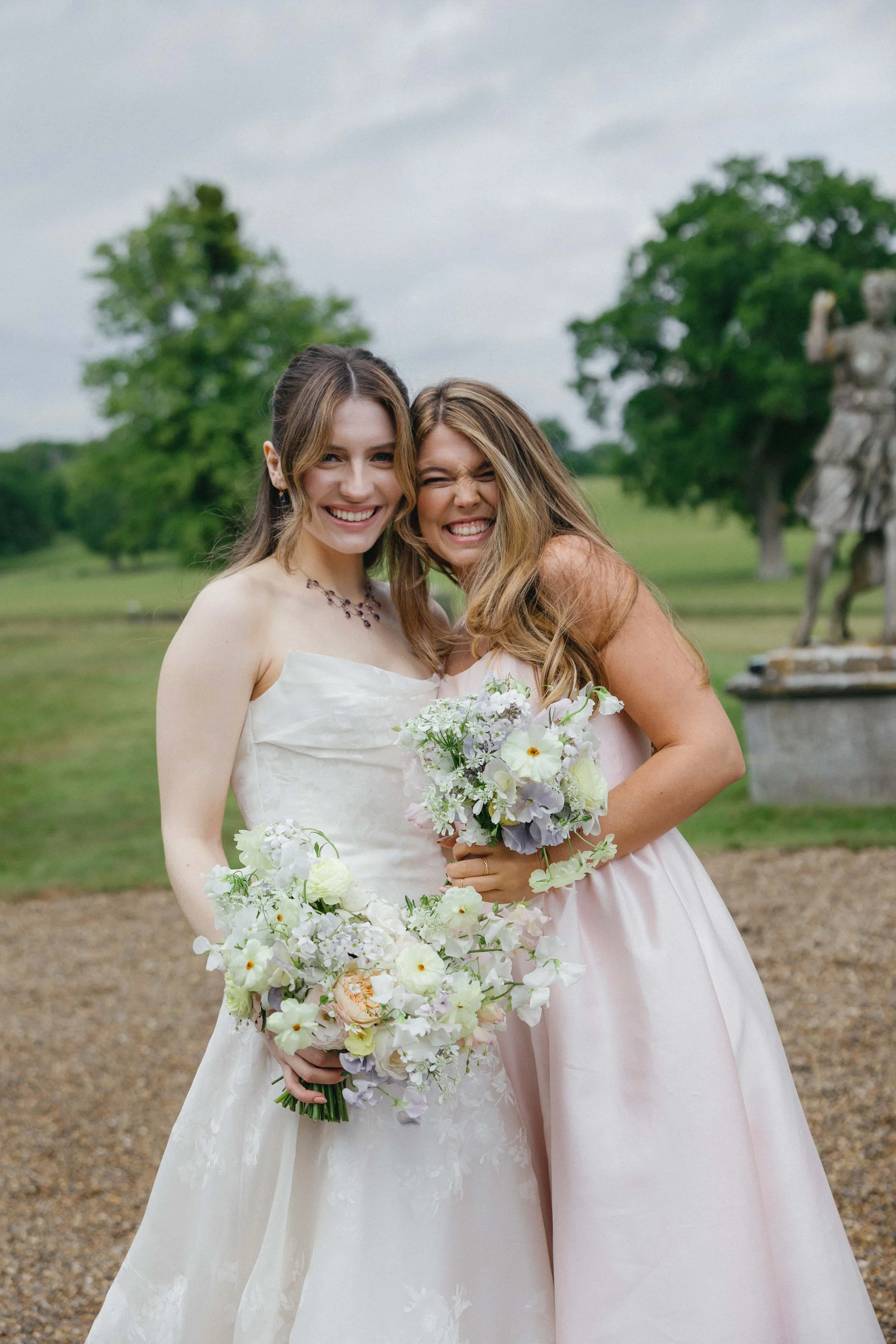 A bride and Bridesmaid holding bouquets, smiling and hugging outdoors with green trees and a monument in the background. Wilderness Reserve, Suffolk Florals By The Botany House
