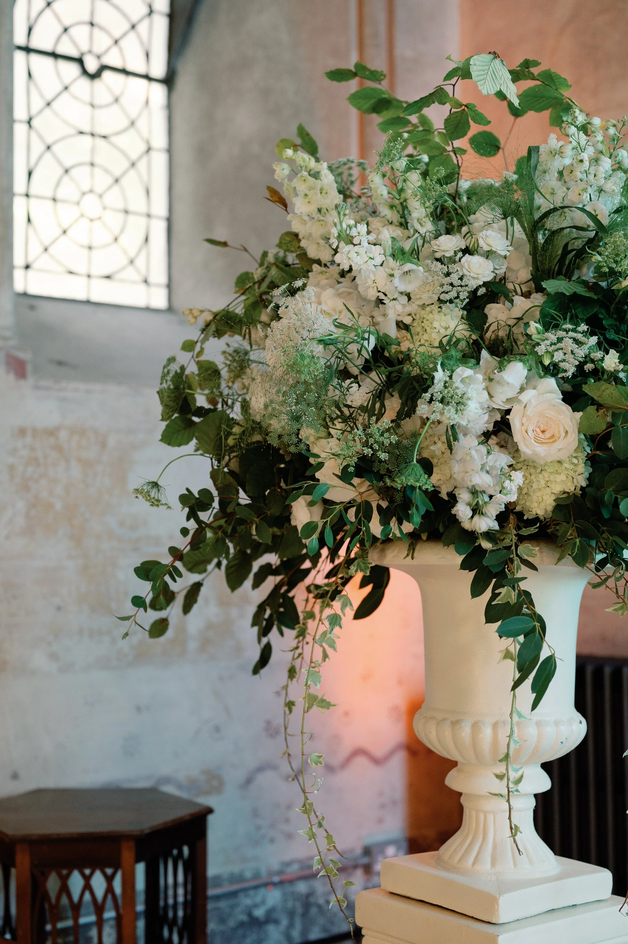 A large elegant floral arrangement with white roses, hydrangeas, and greenery in a white vase, set indoors near a stone wall and a stained glass window.