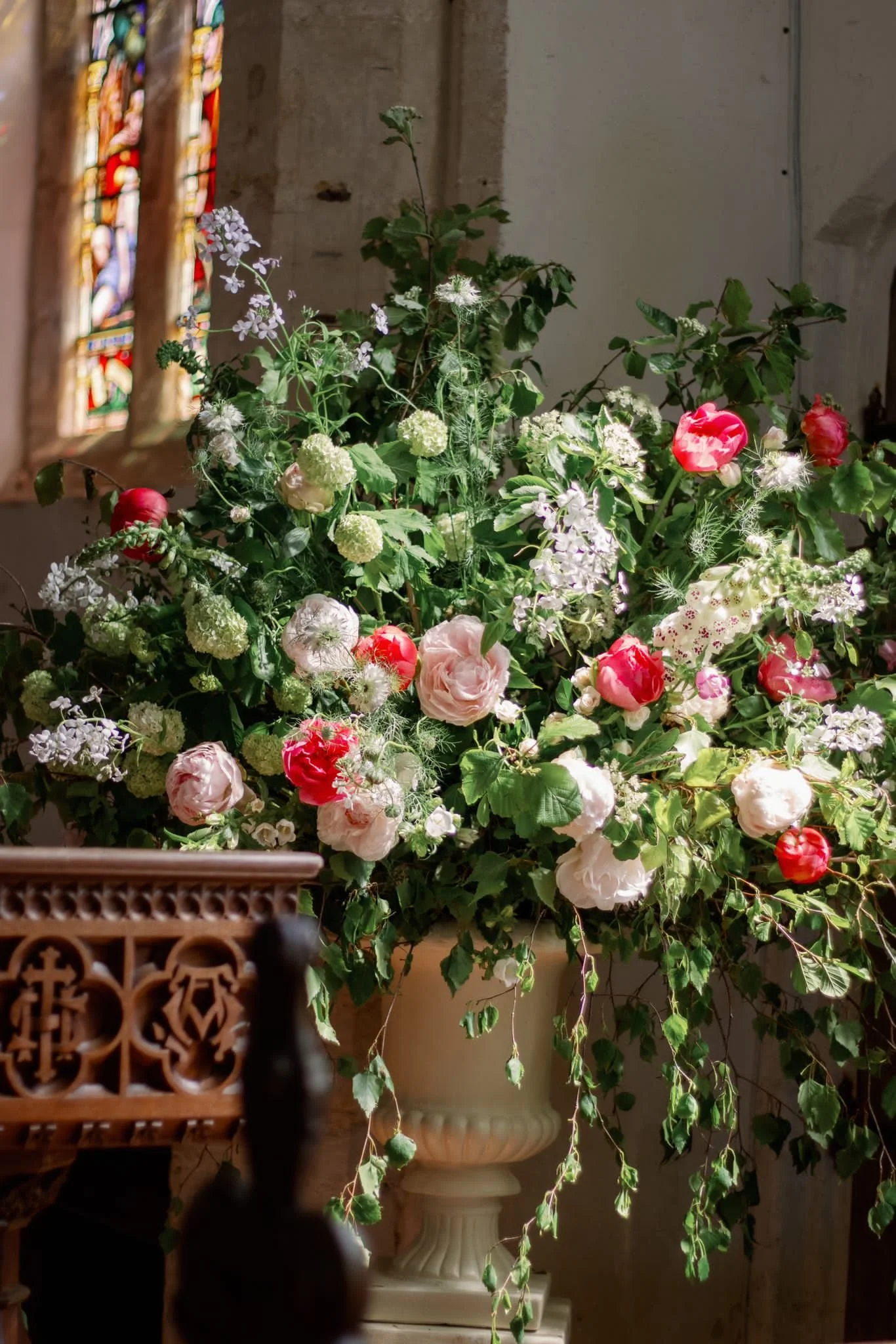 A large arrangement of various blooming flowers inside a church, with a stained-glass window in the background.
