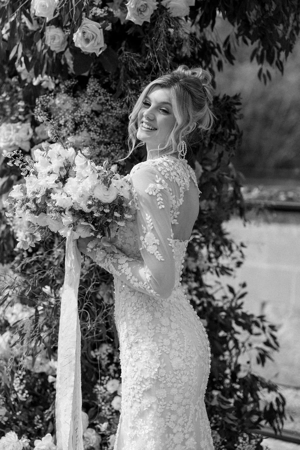 A bride in a lace wedding dress holding a bouquet of flowers, smiling, with flowers and greenery in the background.