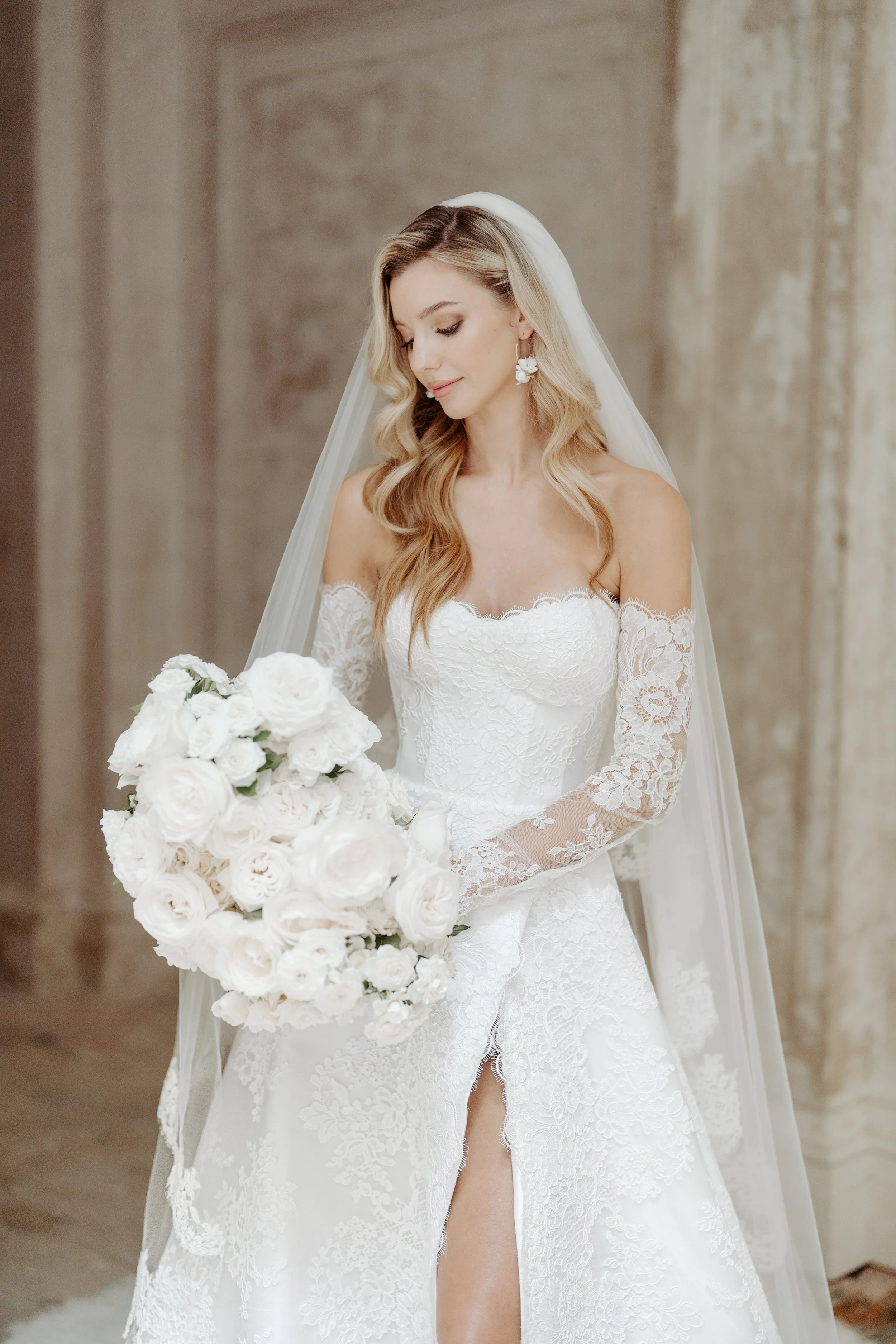 A bride in a white lace wedding gown holding a bouquet of white roses, wearing a veil and floral earrings, standing indoors.