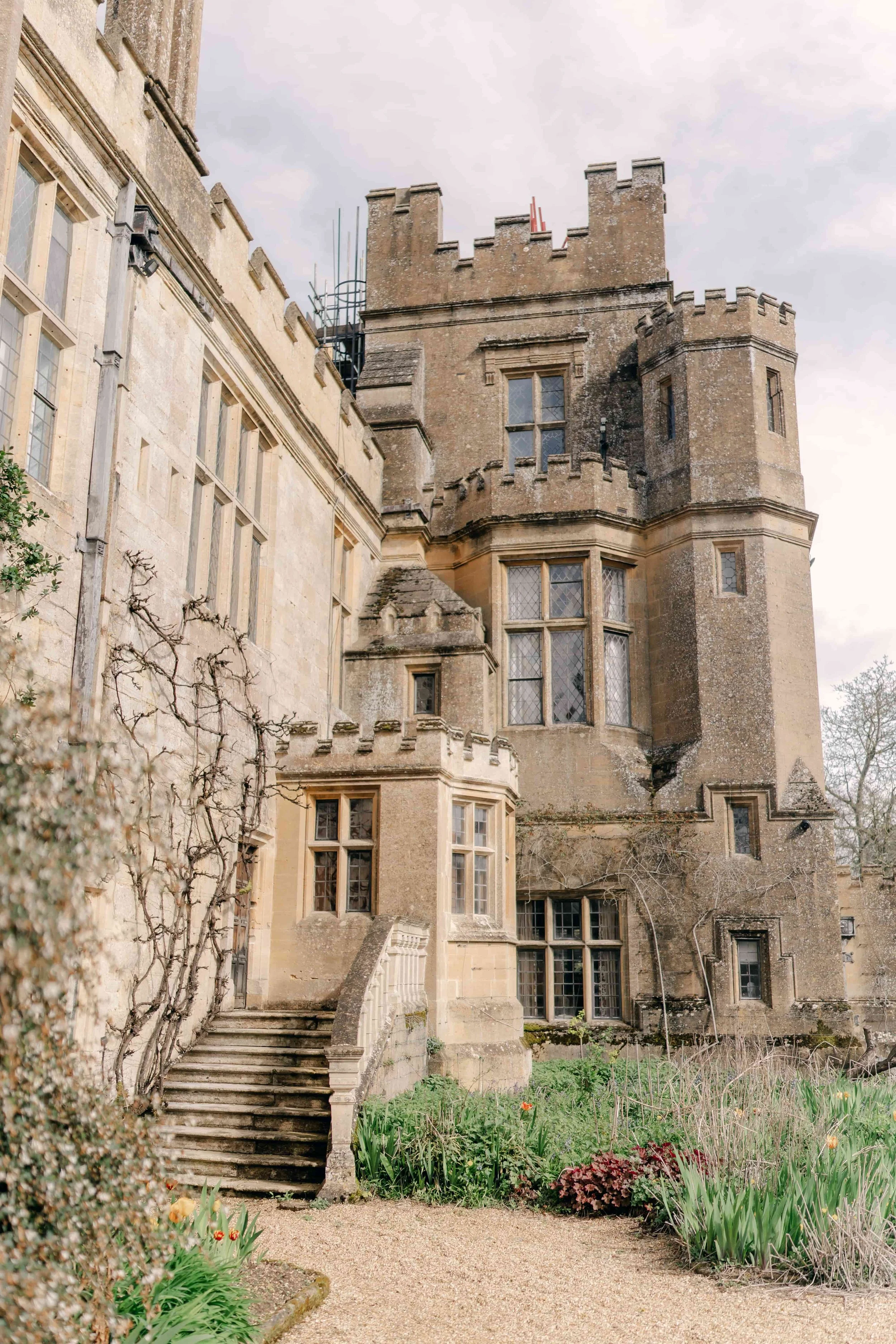 A historic Sudeley castle with stone walls, tower, and gothic windows, surrounded by a garden with plants and flowers in the Cotswolds