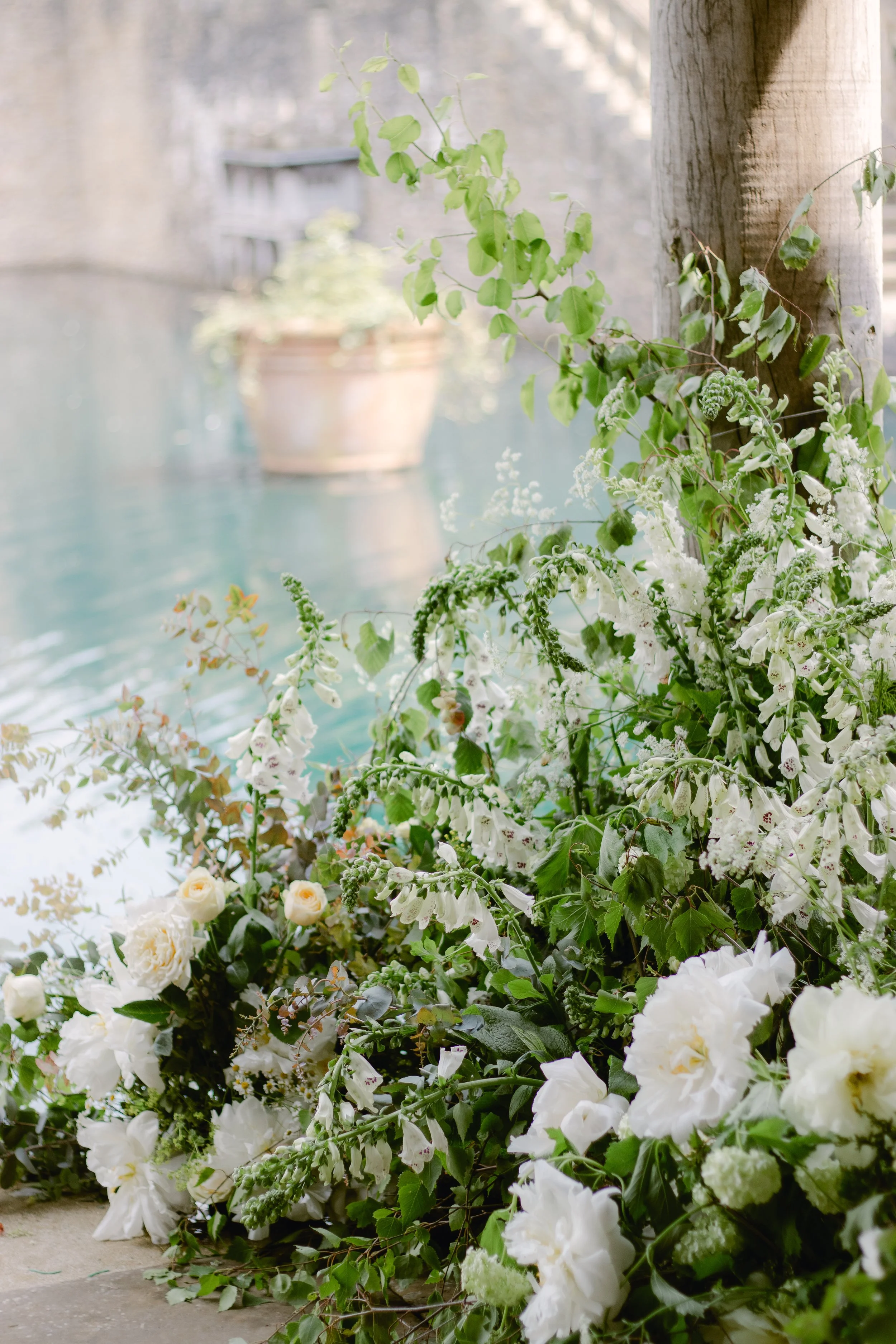 A floral arrangement with white flowers and green leaves near a wooden post, with a blurry background of a pool and a potted plant.