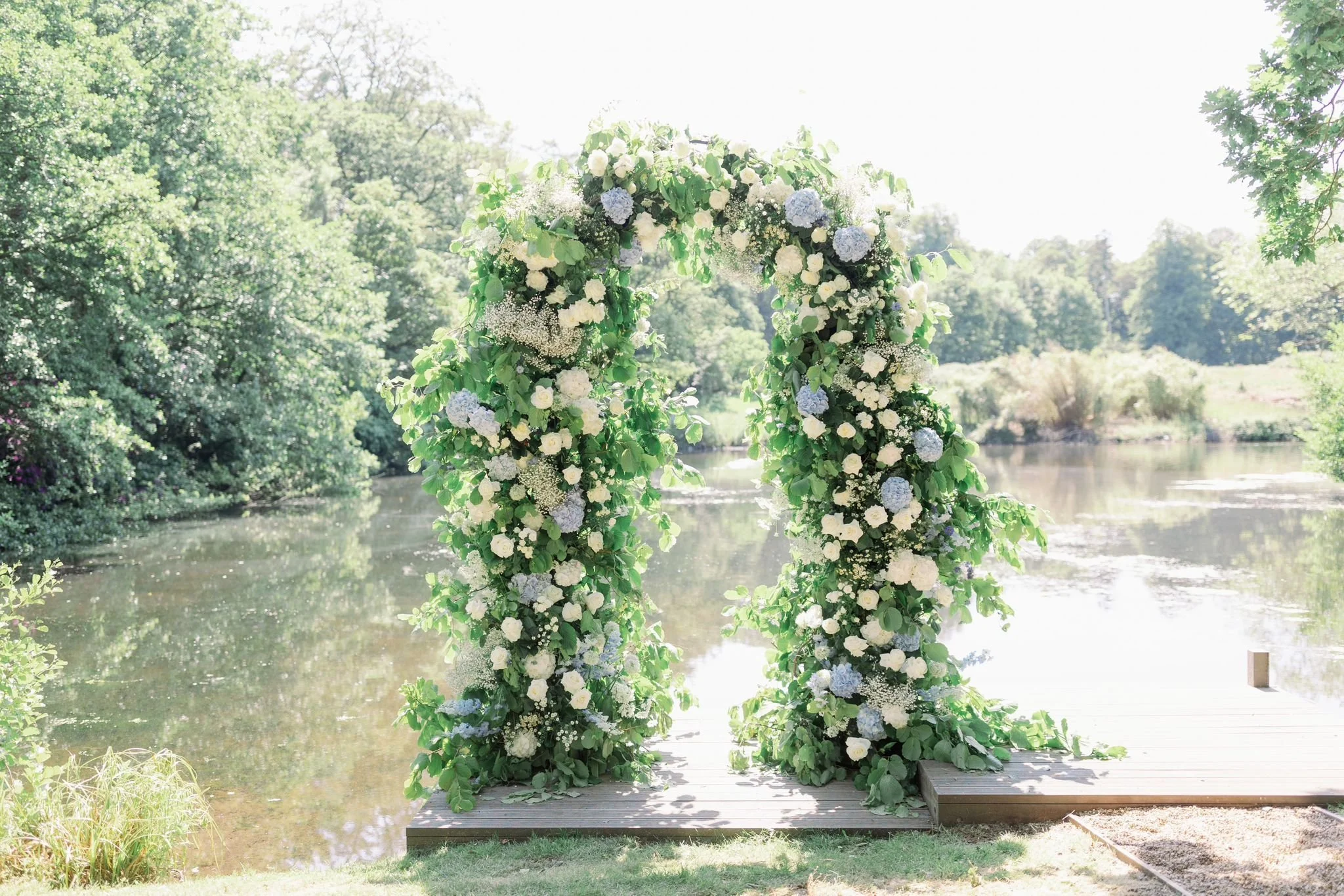 A floral arch decorated with white and light blue flowers and green leaves, set on a wooden platform by a river in a lush, green outdoor setting.