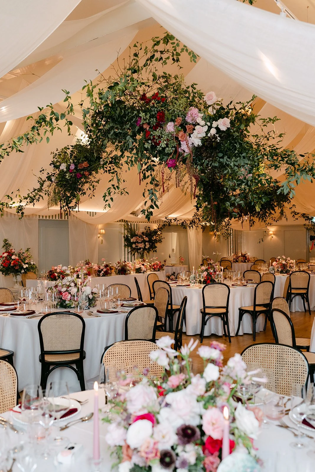 Elegant event setup with round tables covered in white tablecloths, floral centerpieces, candles, and a high floral arrangement hanging from the ceiling in a decorated venue with draped fabric and warm lighting.
